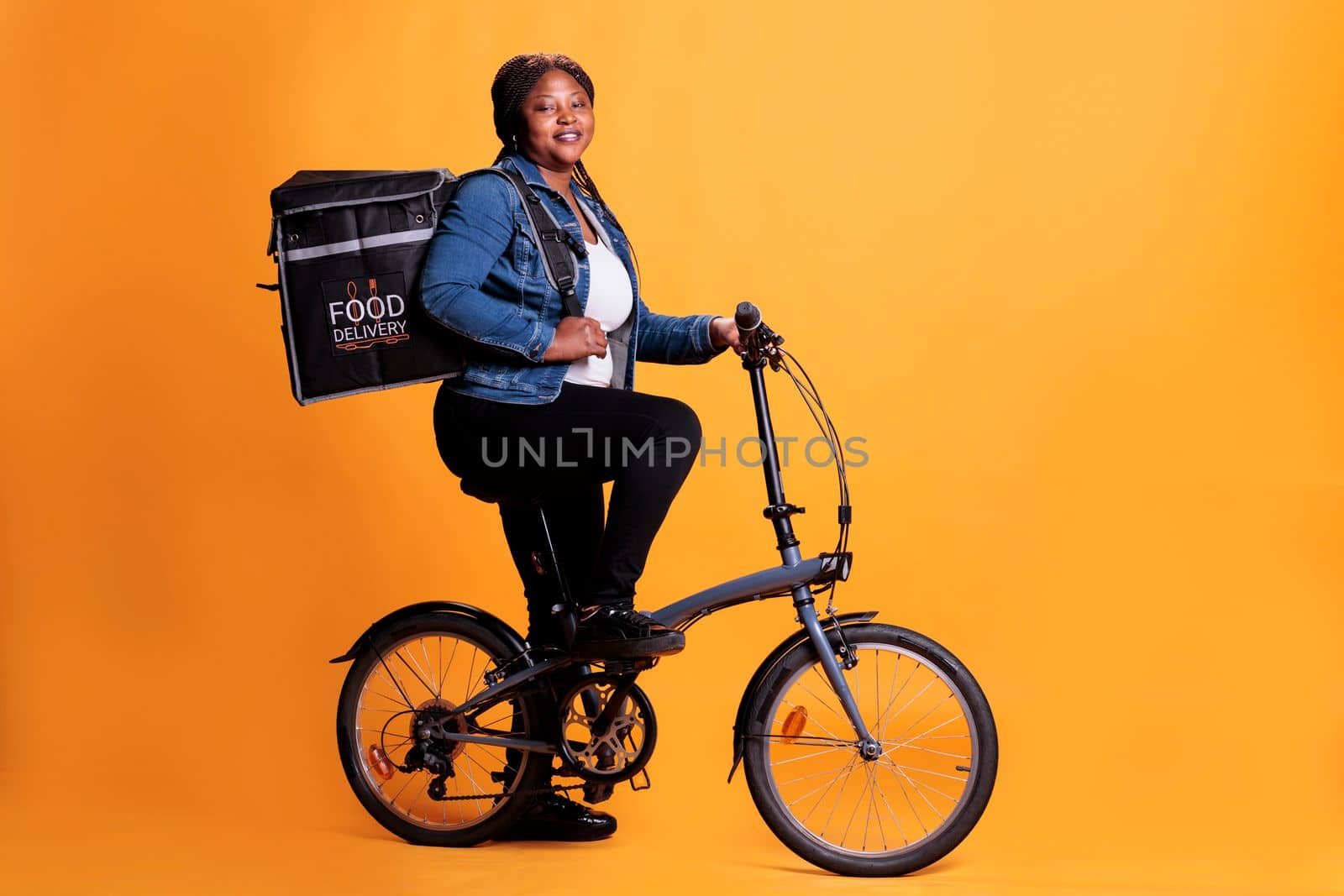 Portrait of fast food delivery worker riding bike during lunch time while carrying thermal takeout backpack by DCStudio