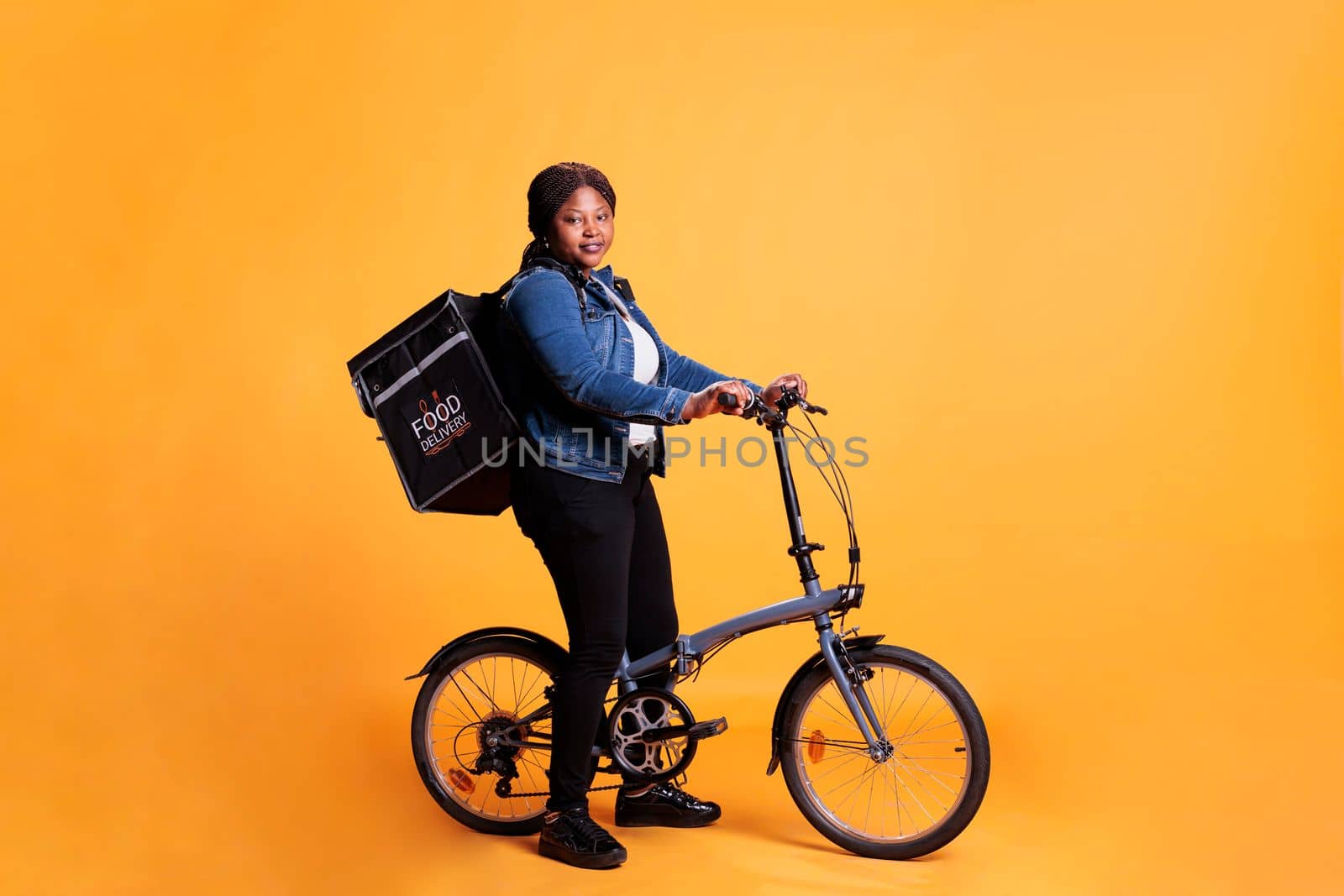 African american woman carrying food delivery thermal backpack standing riding bike in studio with yellow background. Restaurant employee delivering pizza for lunch. Food transportation service