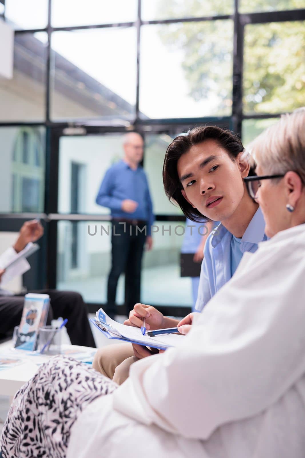 Elderly doctor explaining medication treatment to asian man during checkup visit consultation in hospital waiting area. Young patient holding clipboard filling medical report before examination