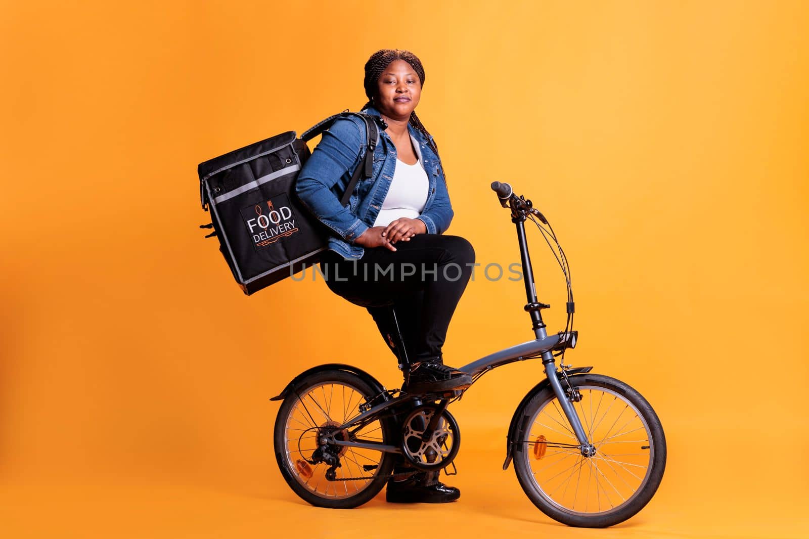 Pizzeria deliverywoman carrying food takeaway backpack while riding bicycle in studio with yellow background, delivering pizza order for dinner. Food transportation service and takeaway concept