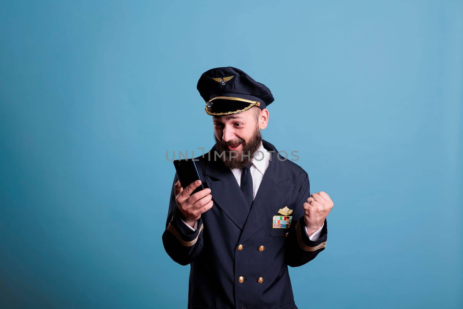 Happy excited airplane captain in uniform holding smartphone with winner gesture, clenched fist. Pilot with cheerful facial expression looking at mobile phone screen, reading good news