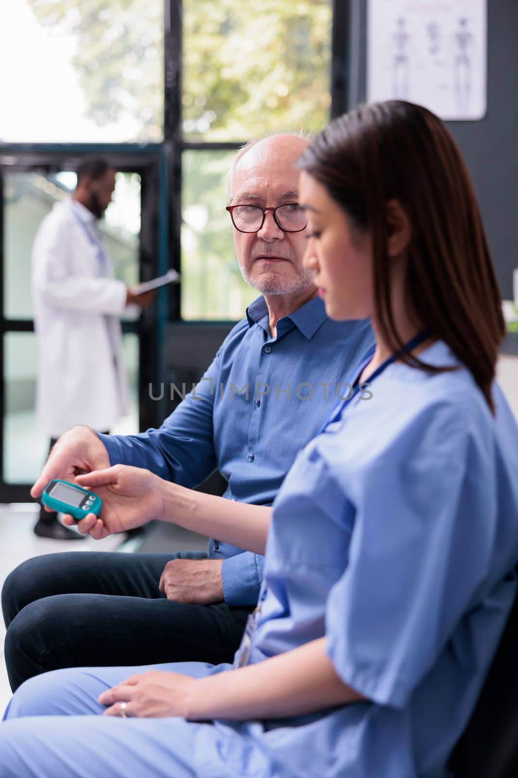 Asian medical asssistant consulting senior patient with glucometer during consultation, measuring insulin and glucose level from blood sample. Old man doing diabetic control in hospital waiting area