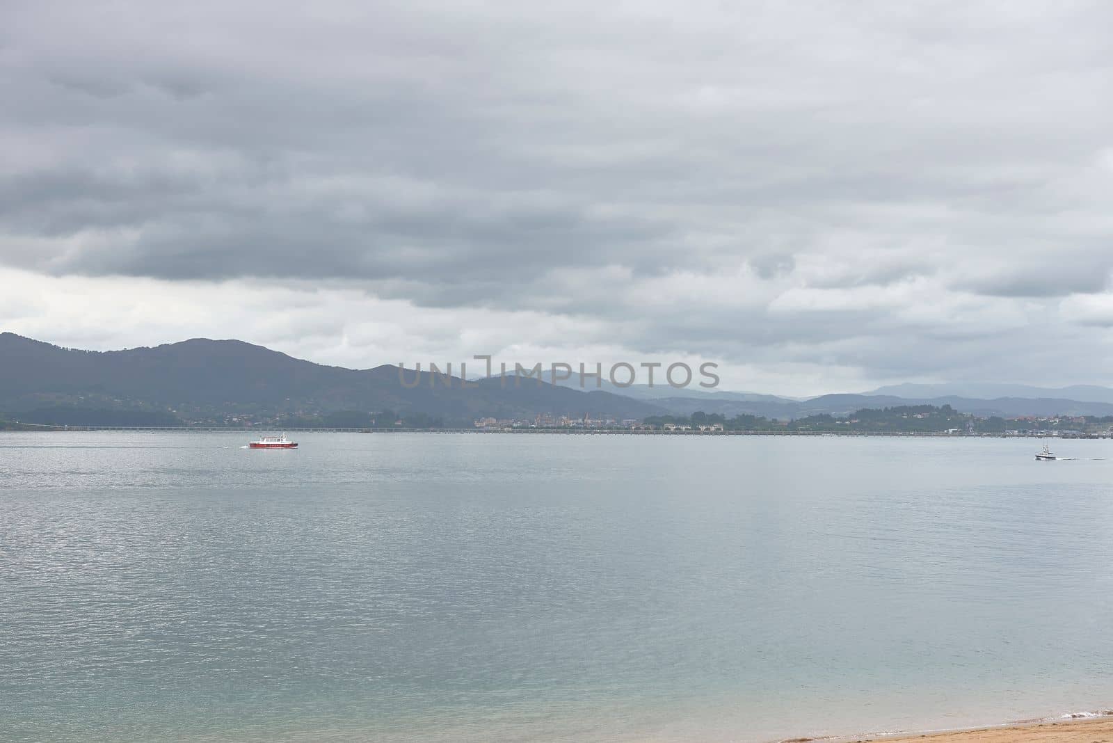 A ship sailing through calm waters in the harbour. Mountains, grey day, empty space,