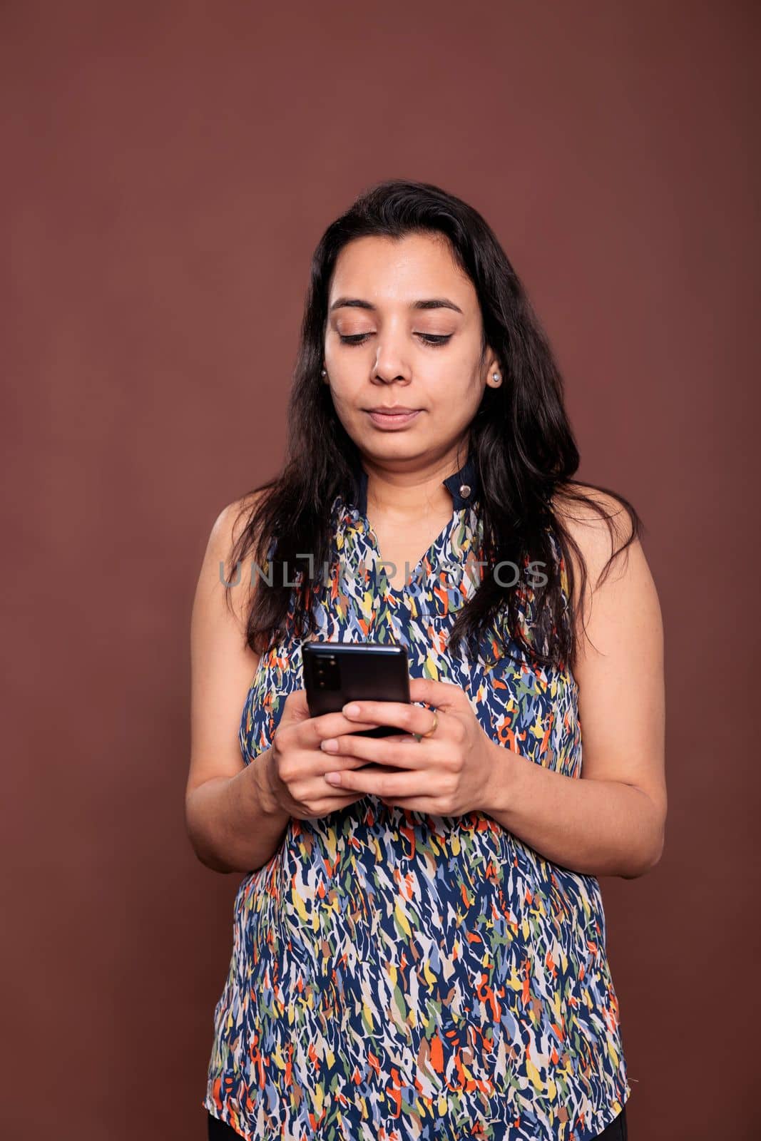 Serious indian woman chatting in social networks, using smartphone, sending sms. Person holding mobile phone, talking online, messaging on telephone, front view studio medium shot