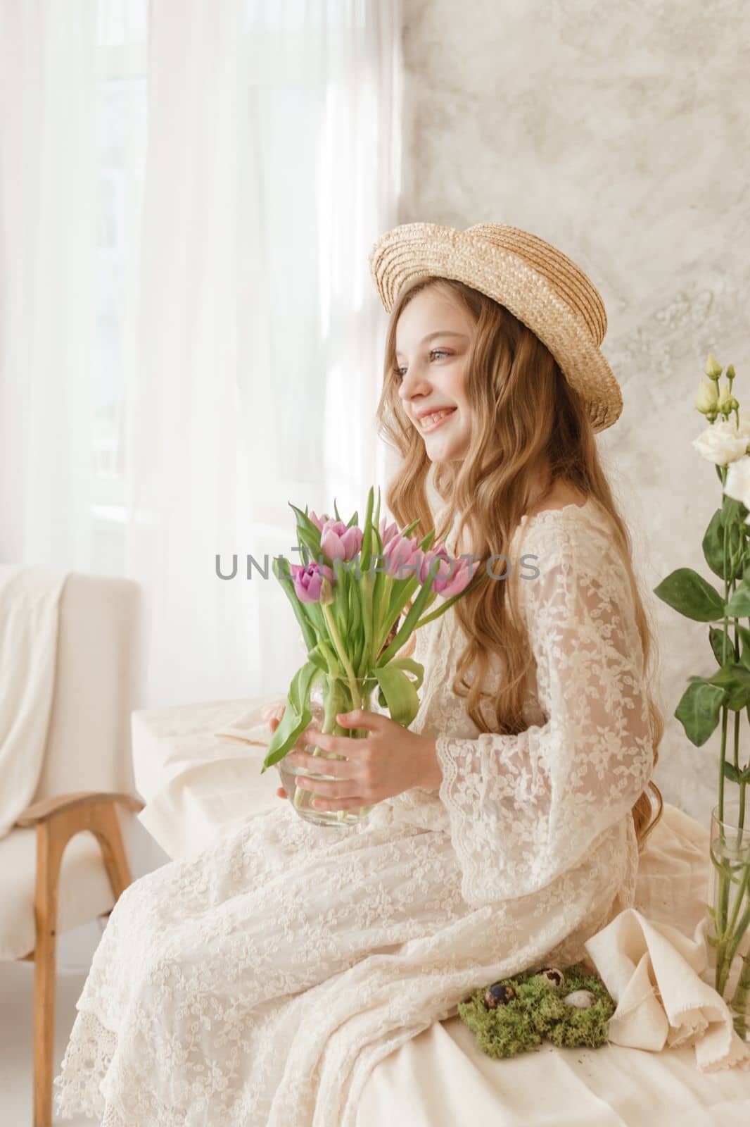 A girl with long hair in a dress and hat is sitting at the Easter table with cakes, spring flowers and quail eggs. Happy Easter celebration