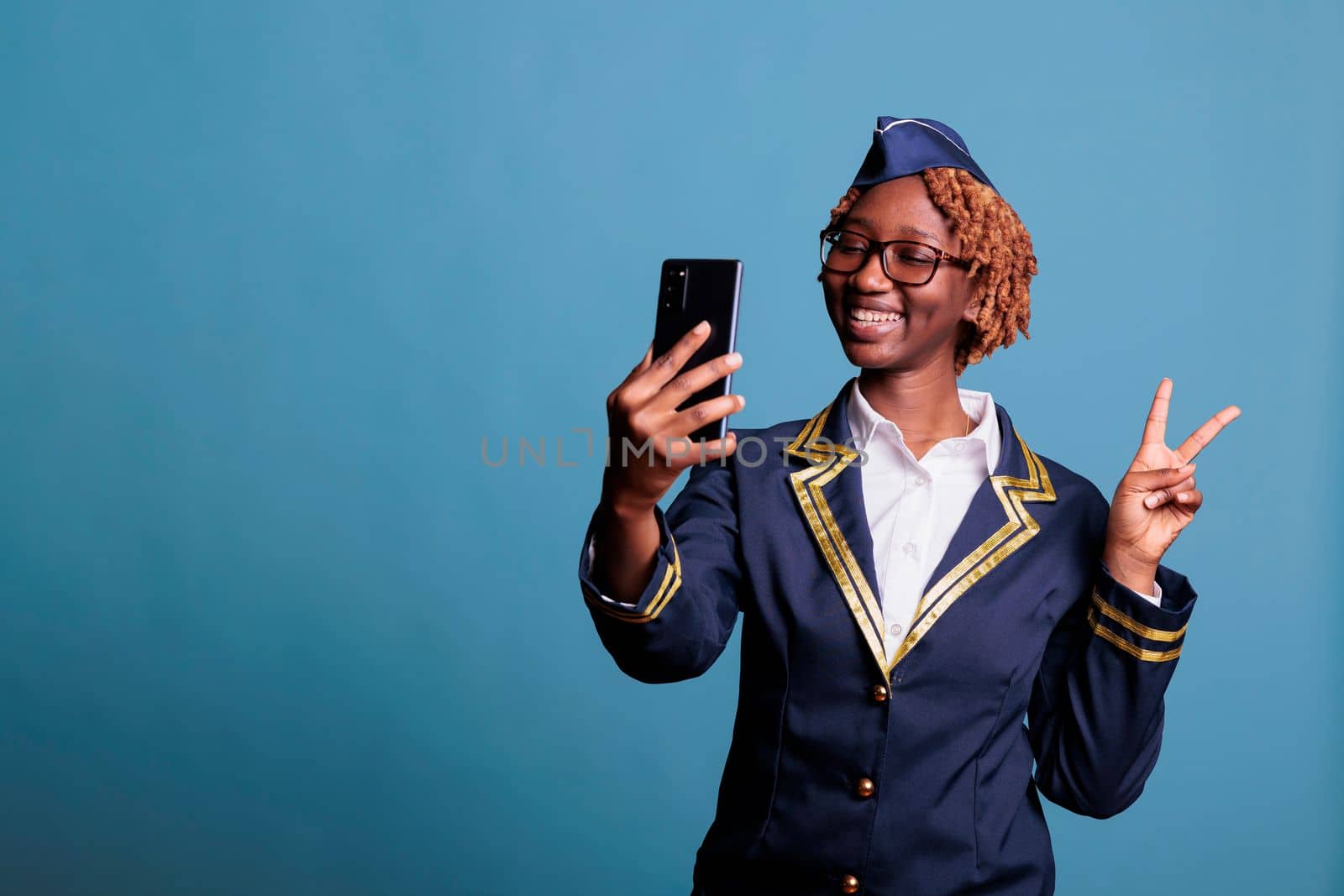 Funny female flight attendant on video call with friends during break time at work. Stewardess dressed in uniform using mobile phone while having fun chatting, studio shot.