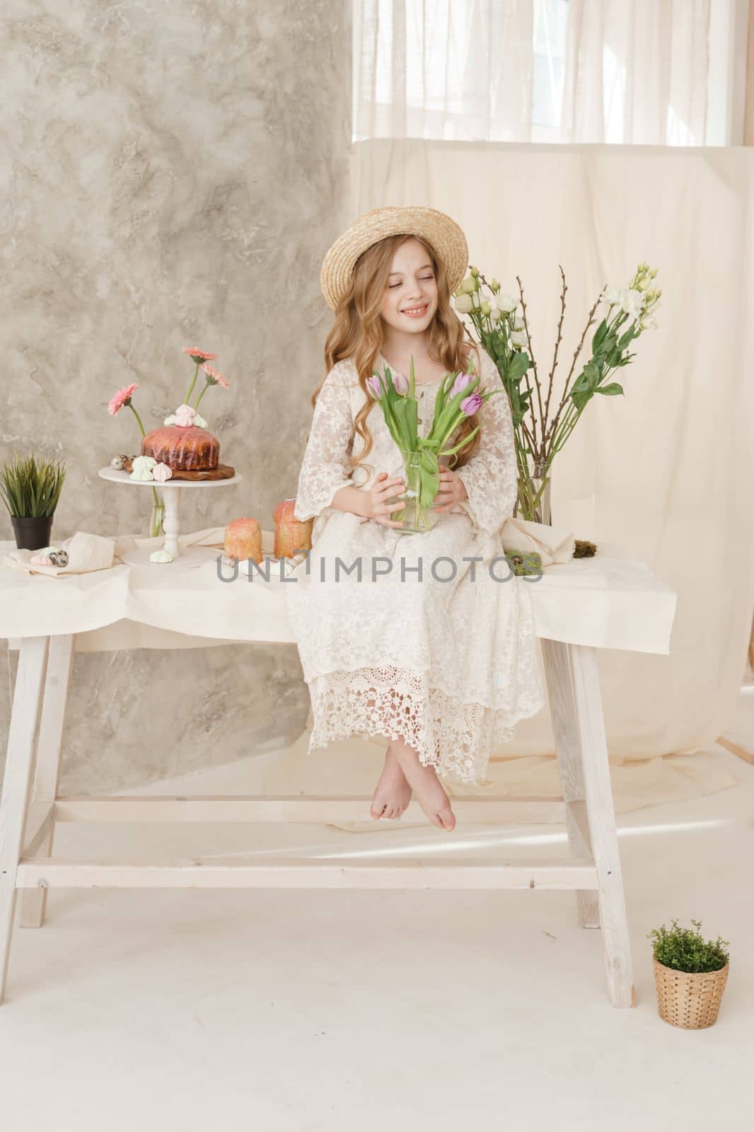 A girl with long hair in a dress and hat is sitting at the Easter table with cakes, spring flowers and quail eggs. Happy Easter celebration