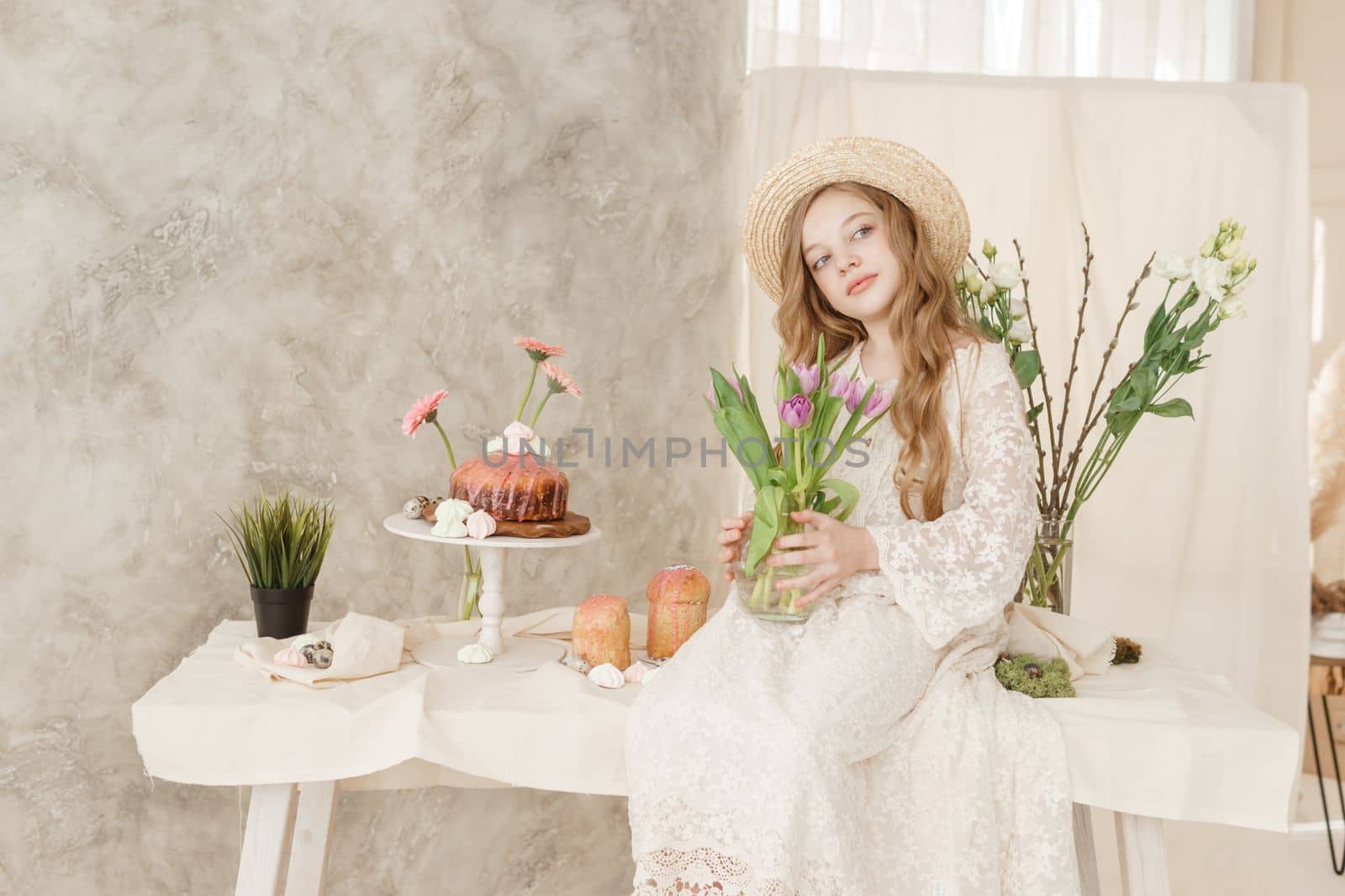 A girl with long hair in a dress and hat is sitting at the Easter table with cakes, spring flowers and quail eggs. Happy Easter celebration