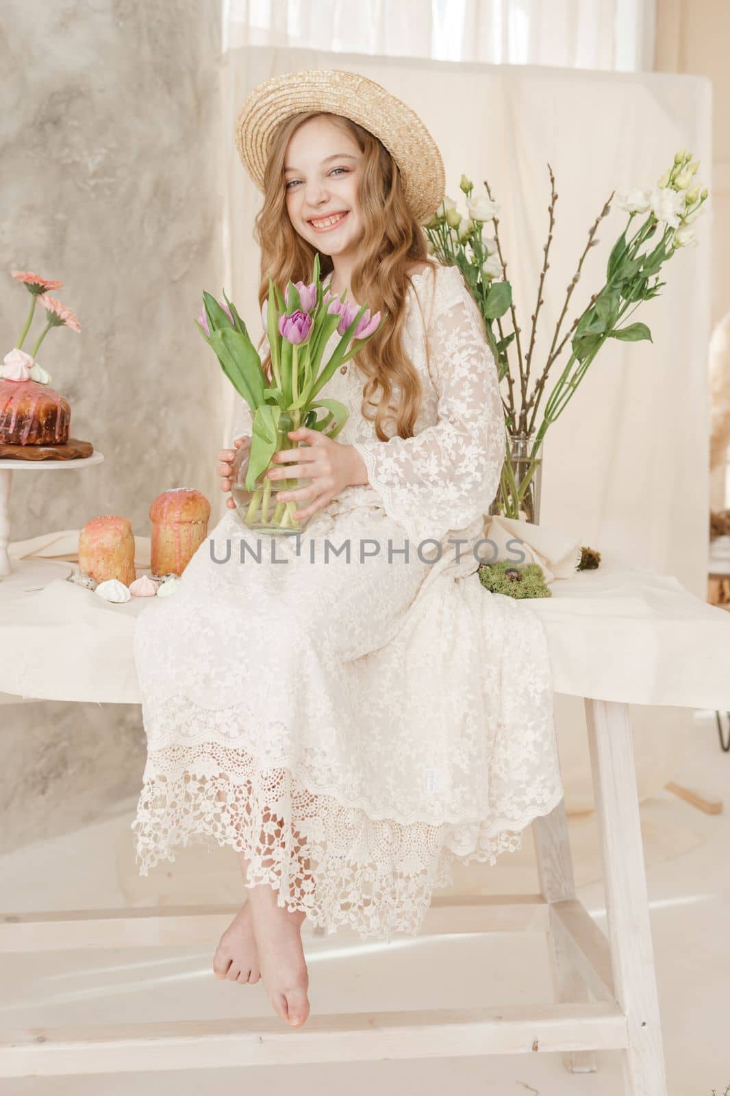 A girl with long hair in a dress and hat is sitting at the Easter table with cakes, spring flowers and quail eggs. Happy Easter celebration