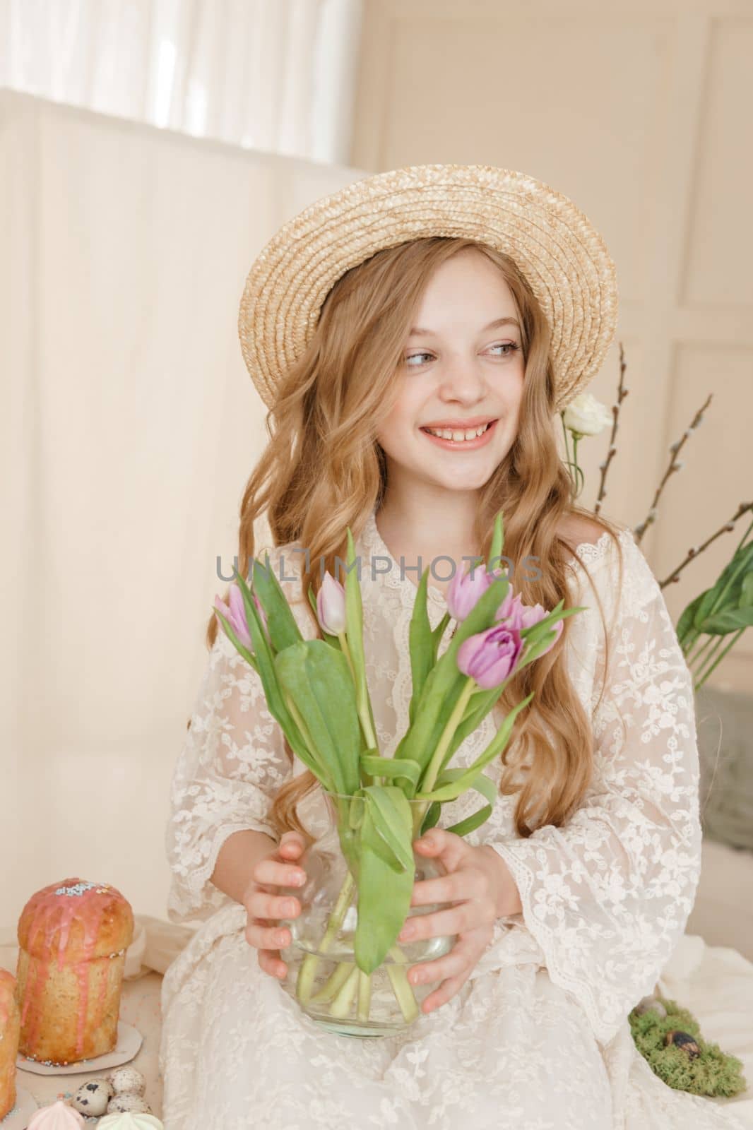 A girl with long hair in a dress and hat is sitting at the Easter table with cakes, spring flowers and quail eggs. Happy Easter celebration
