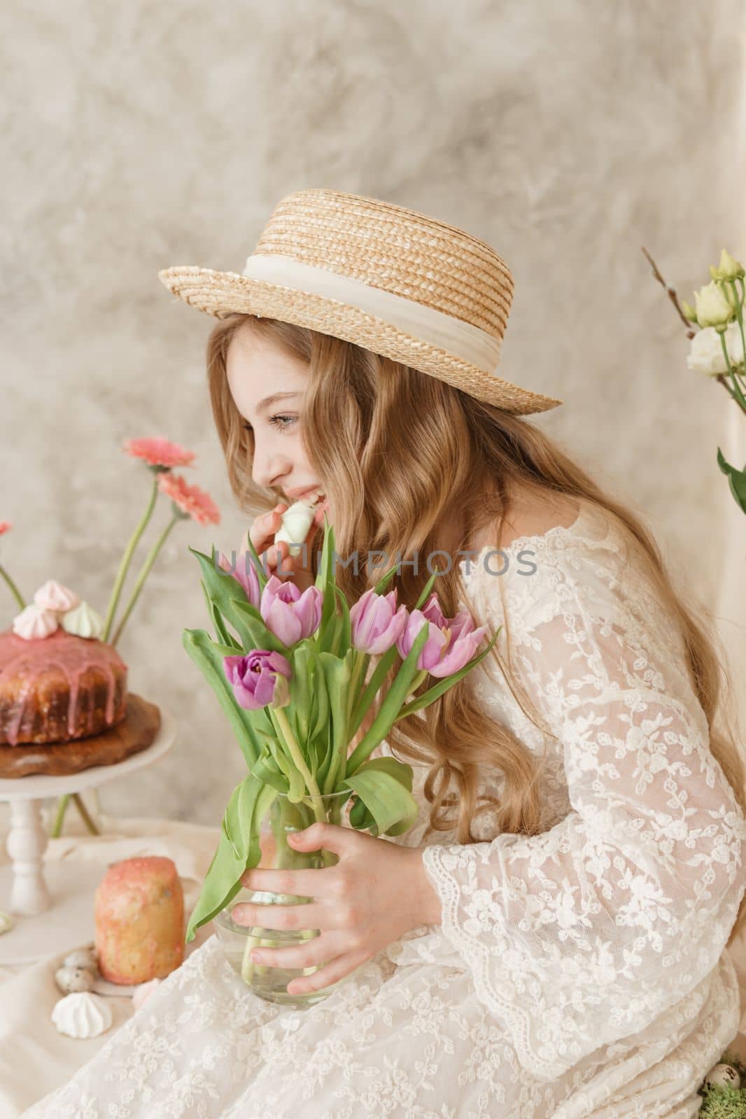 A girl with long hair in a dress and hat is sitting at the Easter table with cakes, spring flowers and quail eggs. Happy Easter celebration