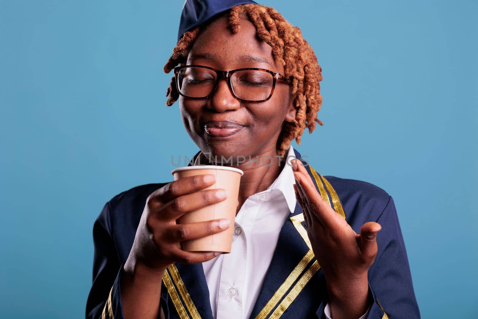 Professional female flight attendant delighted with scent of freshly brewed coffee during break time at work. Air hostess dressed in work uniform against blue background.