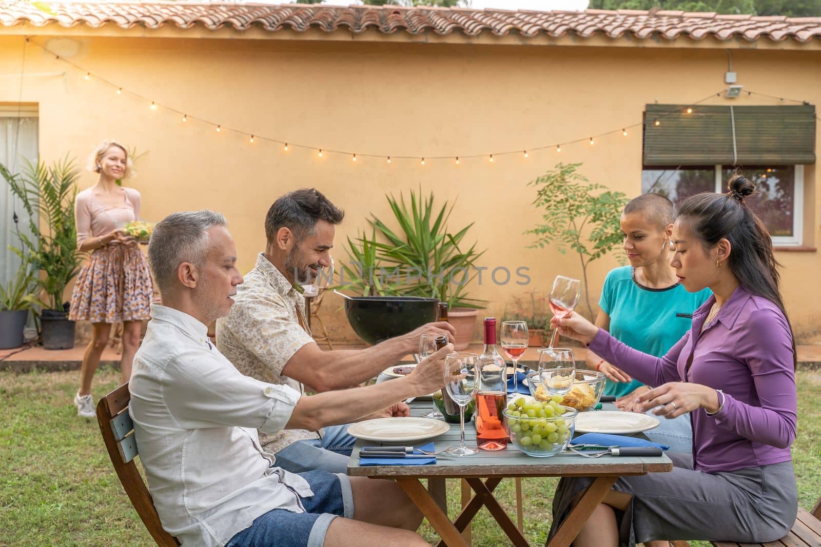 Multicultural happy friends laughing with beautiful smile around the lunch table. While woman brings the food at house patio. High quality photo