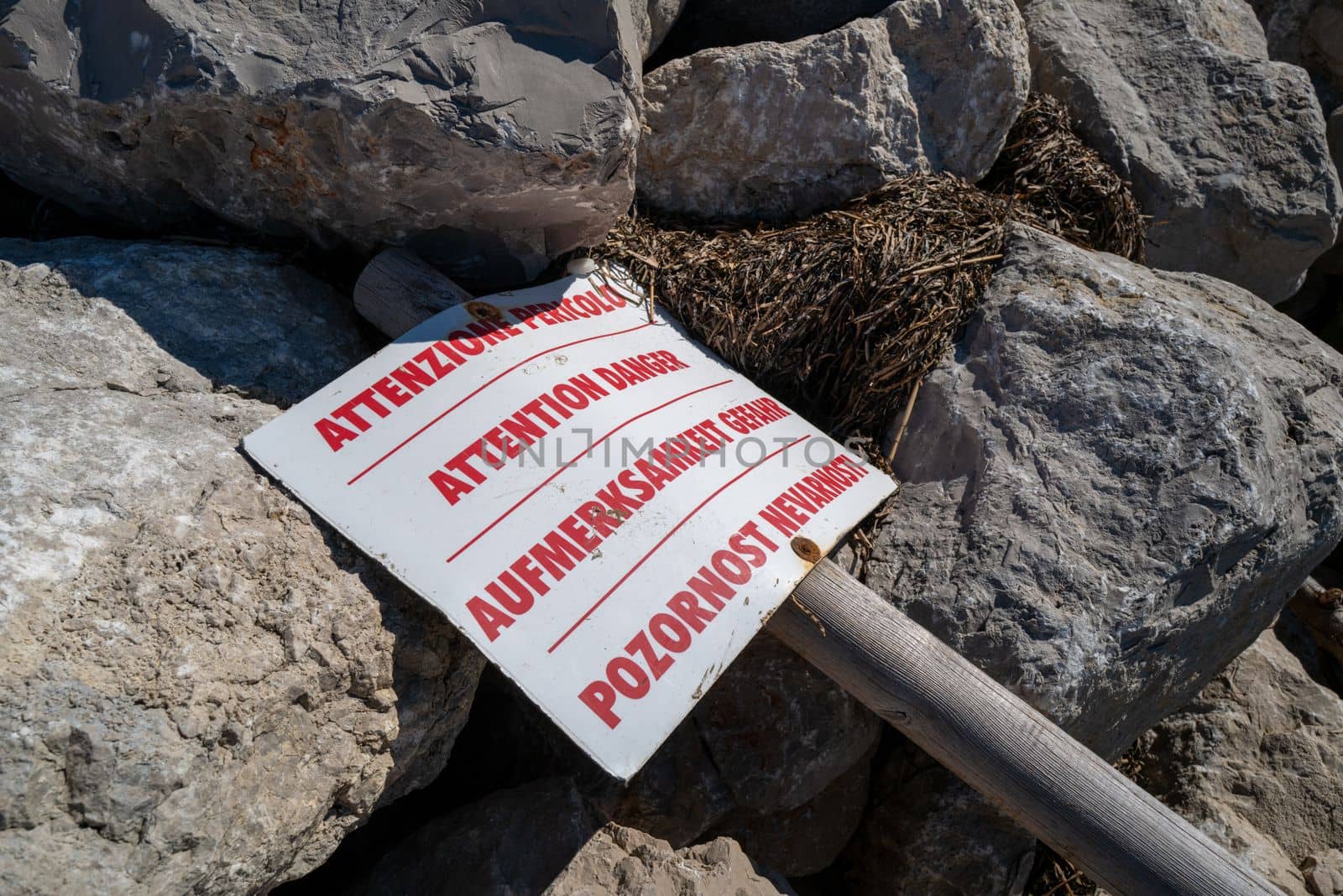 A sign with the inscription "Warning" in four different languages lies on a pile of large stones.