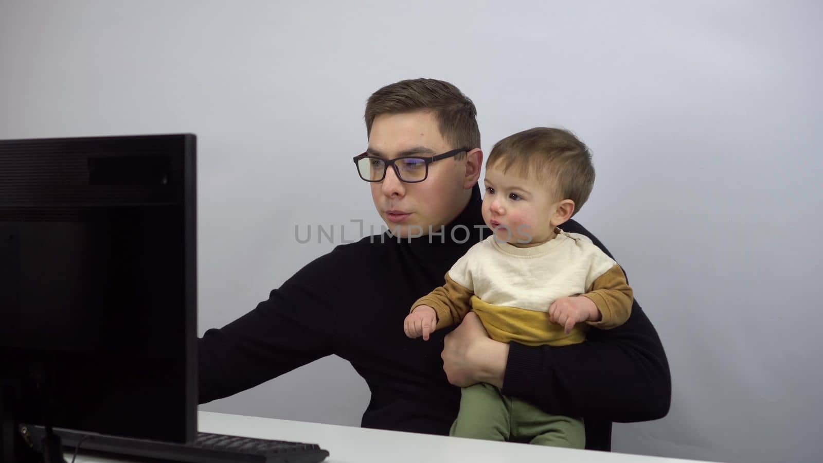 A young IT specialist works at a computer with a small son. A woman gives her son into the hands of a husband who works remotely from home. A man with glasses and a black turtleneck. 4k