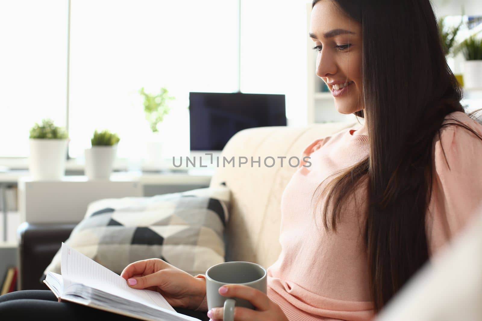 Smiling woman is reading a book and holding cup of tea on sofa. Home leisure and hobby concept