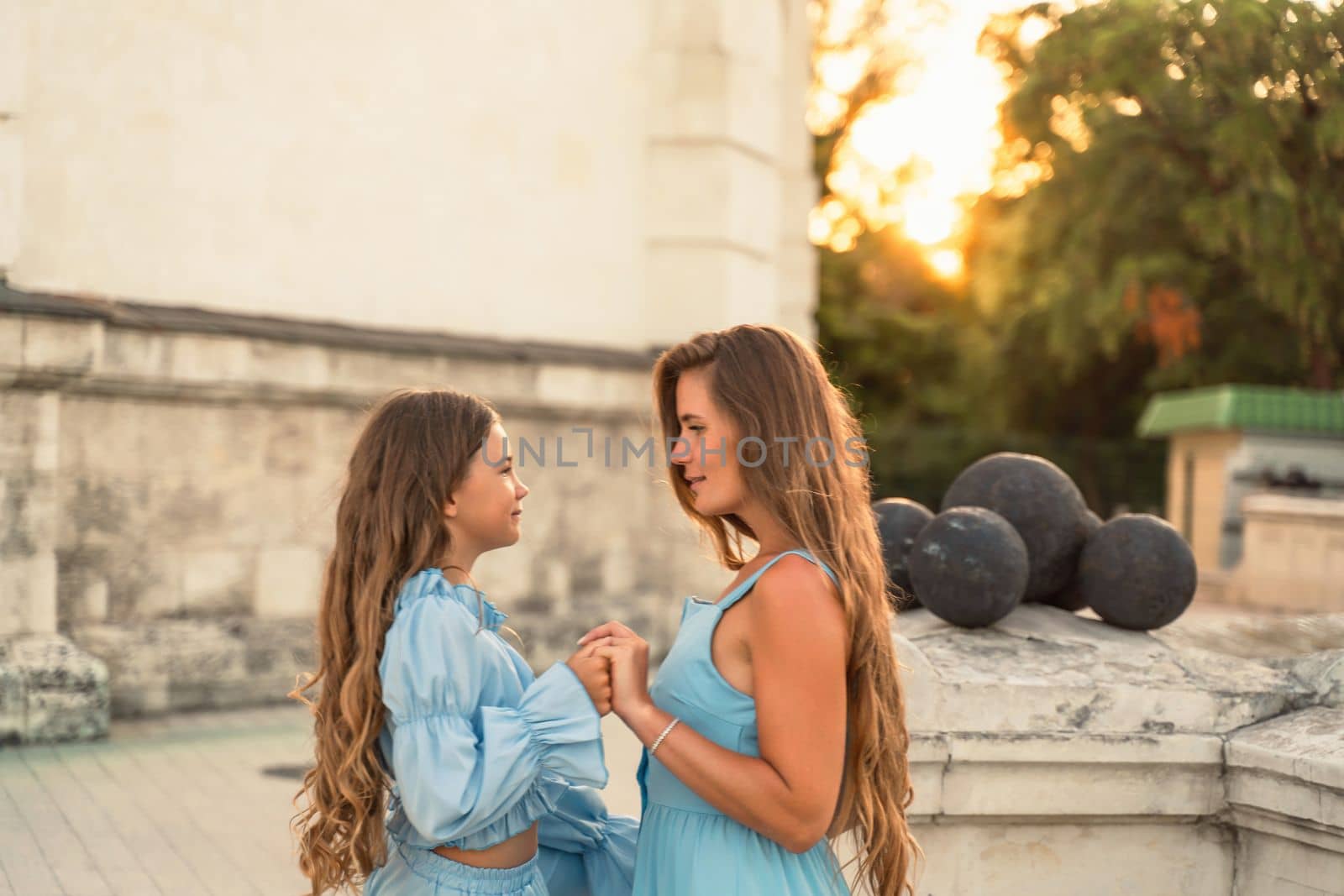 Portrait of a mother and daughter in blue dresses with flowing long hair against the backdrop of a sunset and a white building. They look at each other. Family stories on the weekend by Matiunina