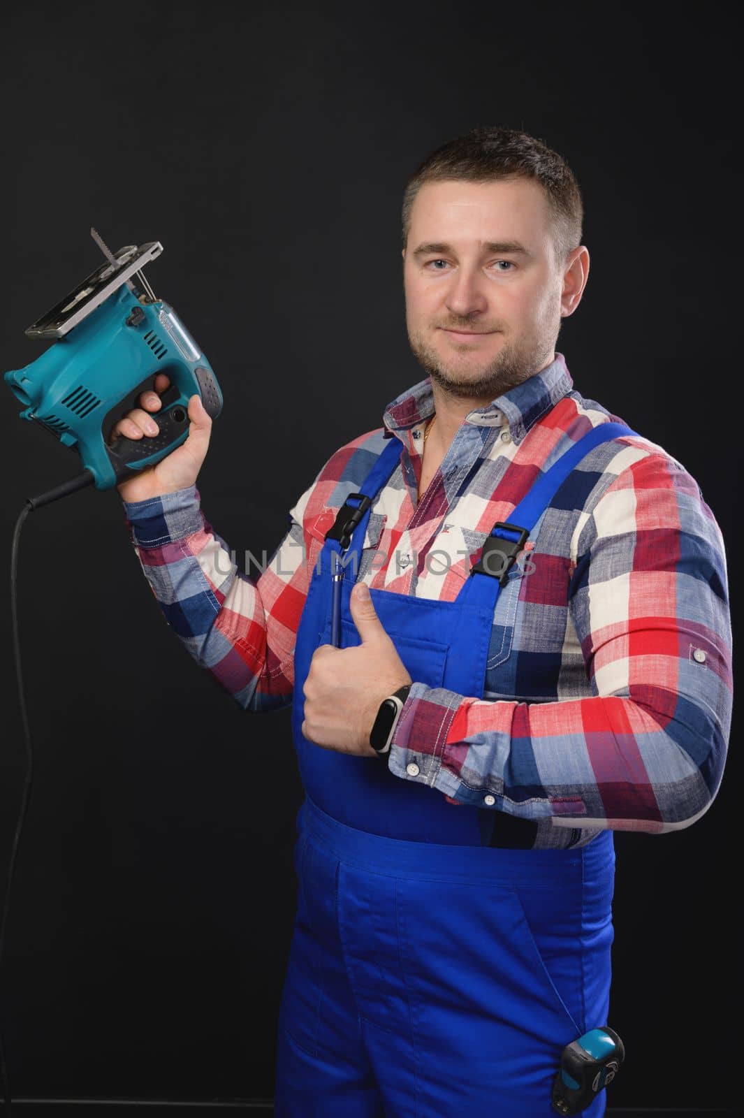Smiling man in a plaid shirt and overalls holds a jigsaw in his hand. on a black background and with the other hand shows a thumbs up gesture.