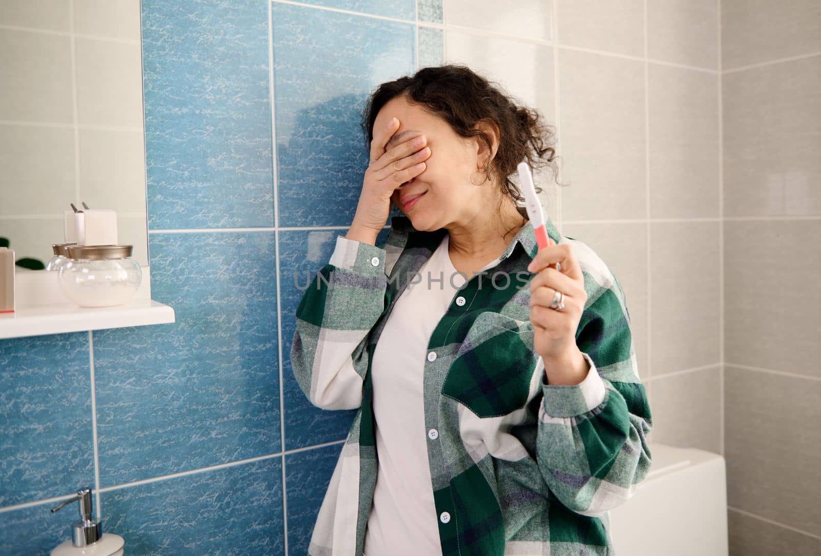 Middle-aged multi-ethnic woman covering her face, surprised by the positive pregnancy test. Pregnant woman showing the test at camera, leaning on the wall of home bathroom