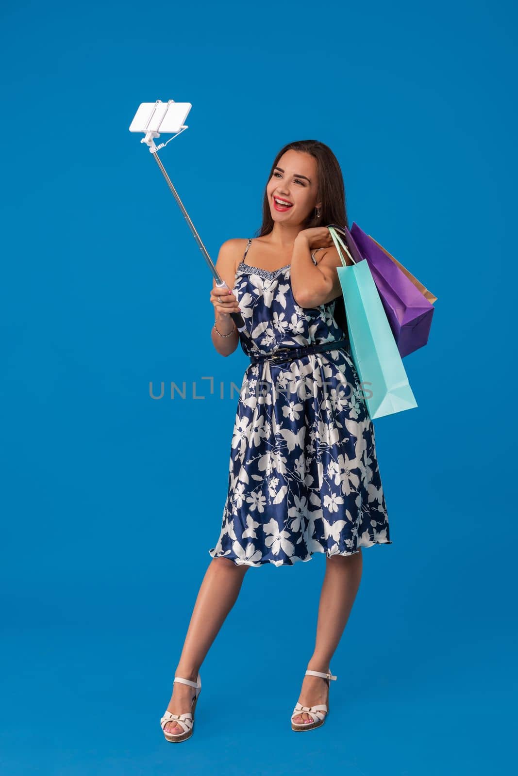 Trendy cheerful woman in casual clothes with shopping bags using selfie stick to take a self portrait on blue studio background with copy space. Emotions. Emotional woman