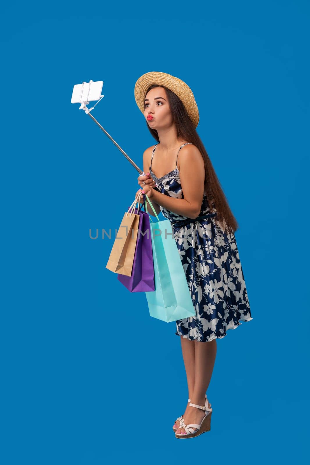 Young woman in casual clothes with shopping bags using selfie stick to take a self portrait on blue studio background with copy space by nazarovsergey