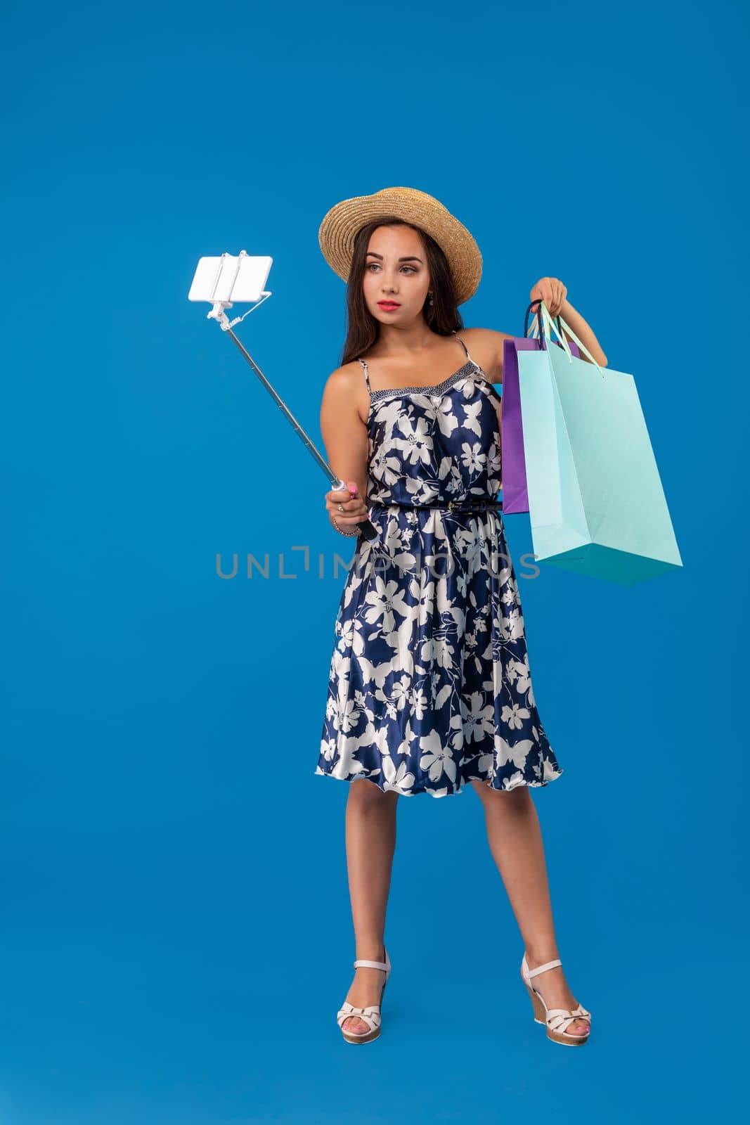 Trendy cheerful woman in casual clothes with shopping bags using selfie stick to take a self portrait on blue studio background with copy space. Emotions. Emotional woman