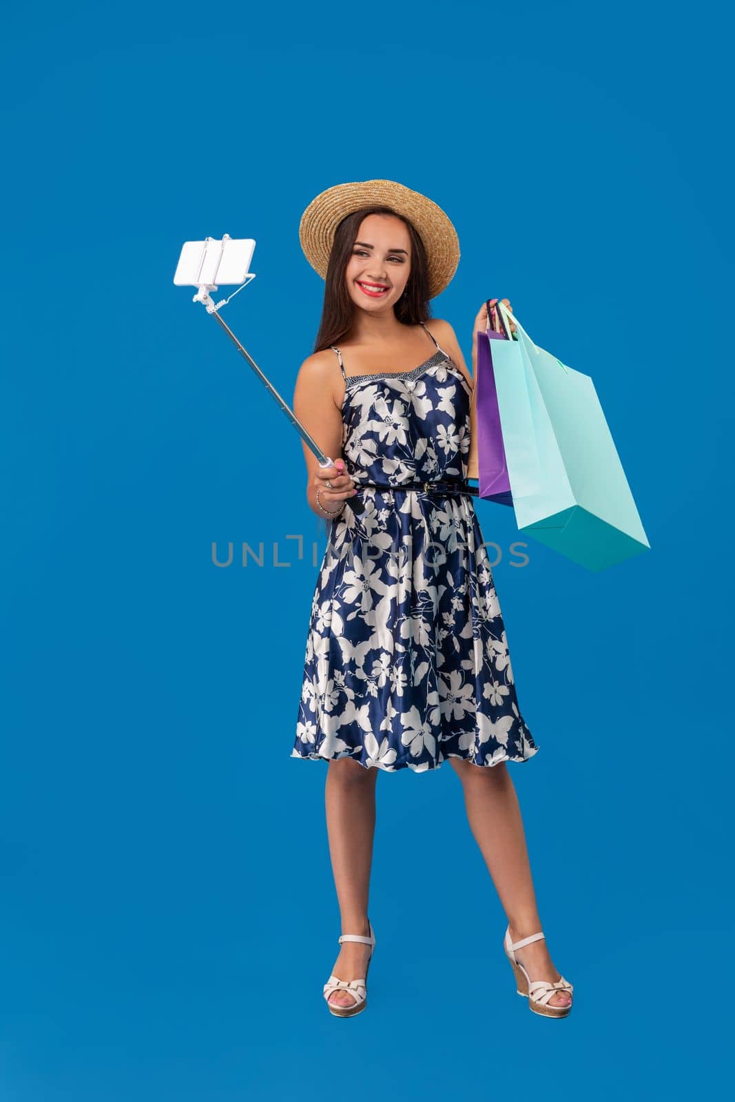 Young woman in casual clothes with shopping bags using selfie stick to take a self portrait on blue studio background with copy space by nazarovsergey