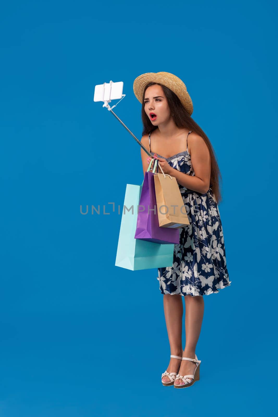 Young woman in casual clothes with shopping bags using selfie stick to take a self portrait on blue studio background with copy space by nazarovsergey