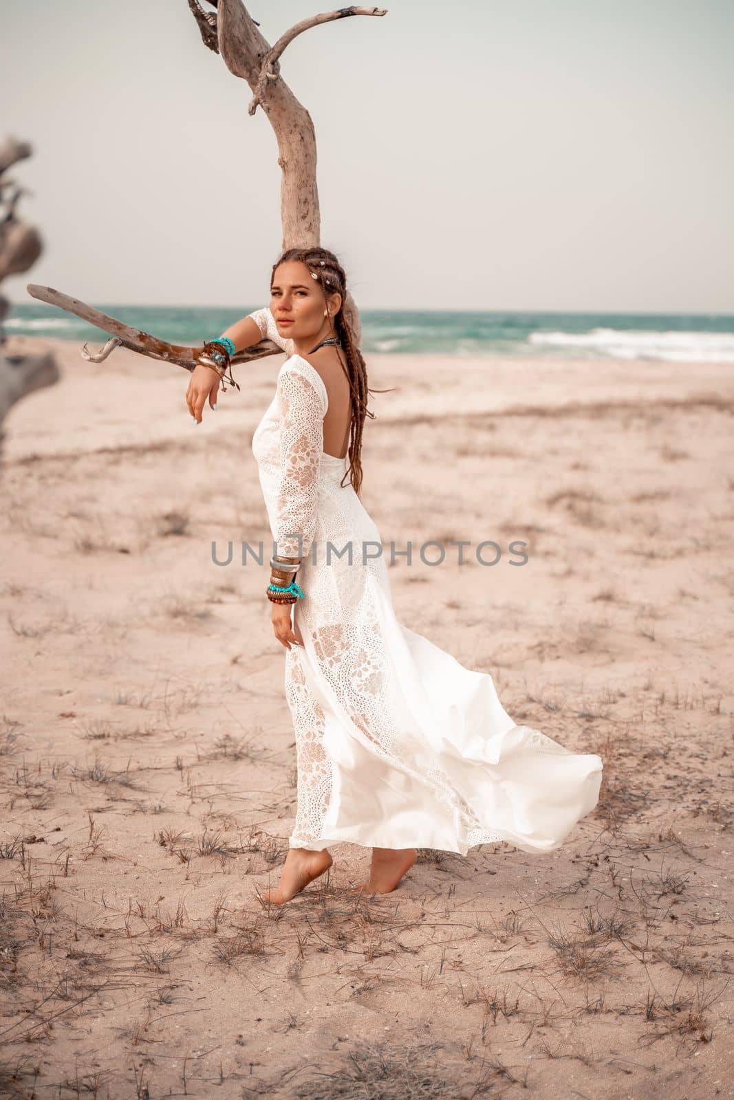 Model in boho style in a white long dress and silver jewelry on the beach. Her hair is braided, and there are many bracelets on her arms