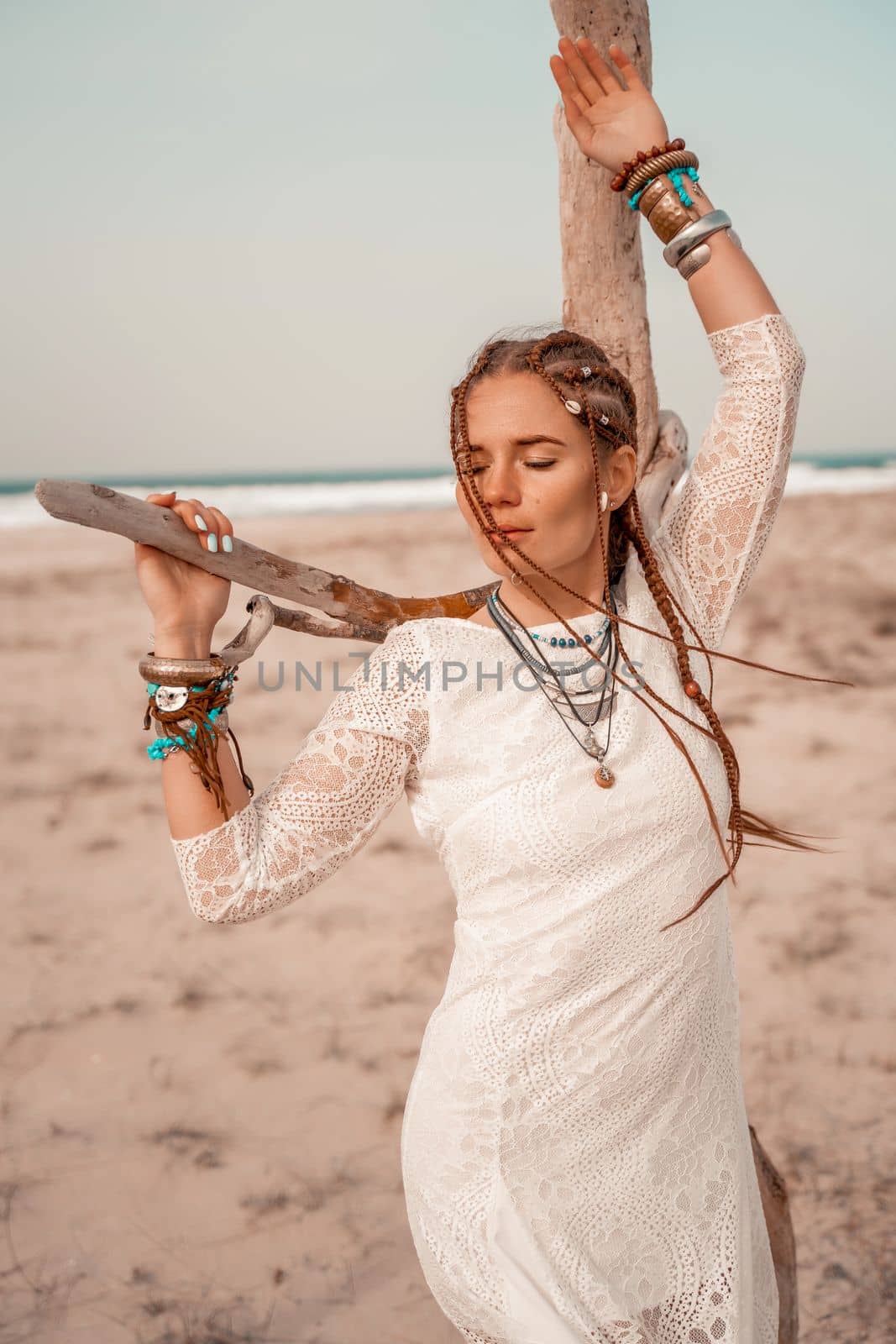 Model in boho style in a white long dress and silver jewelry on the beach. Her hair is braided, and there are many bracelets on her arms