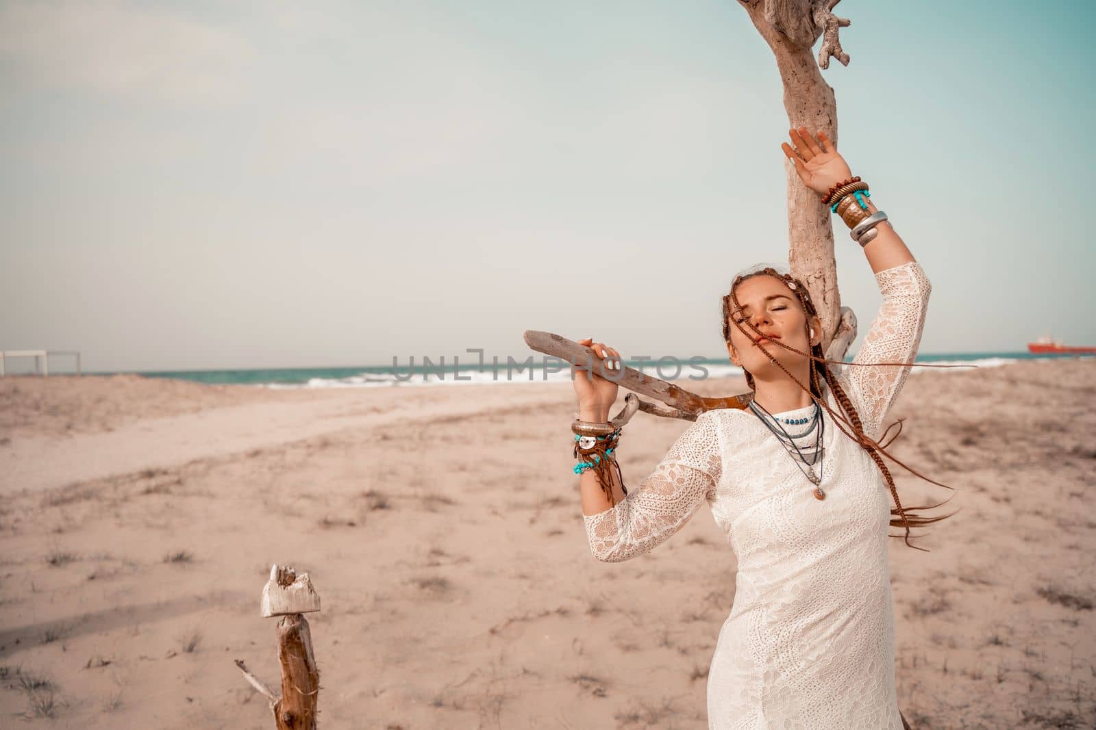 Model in boho style in a white long dress and silver jewelry on the beach. Her hair is braided, and there are many bracelets on her arms