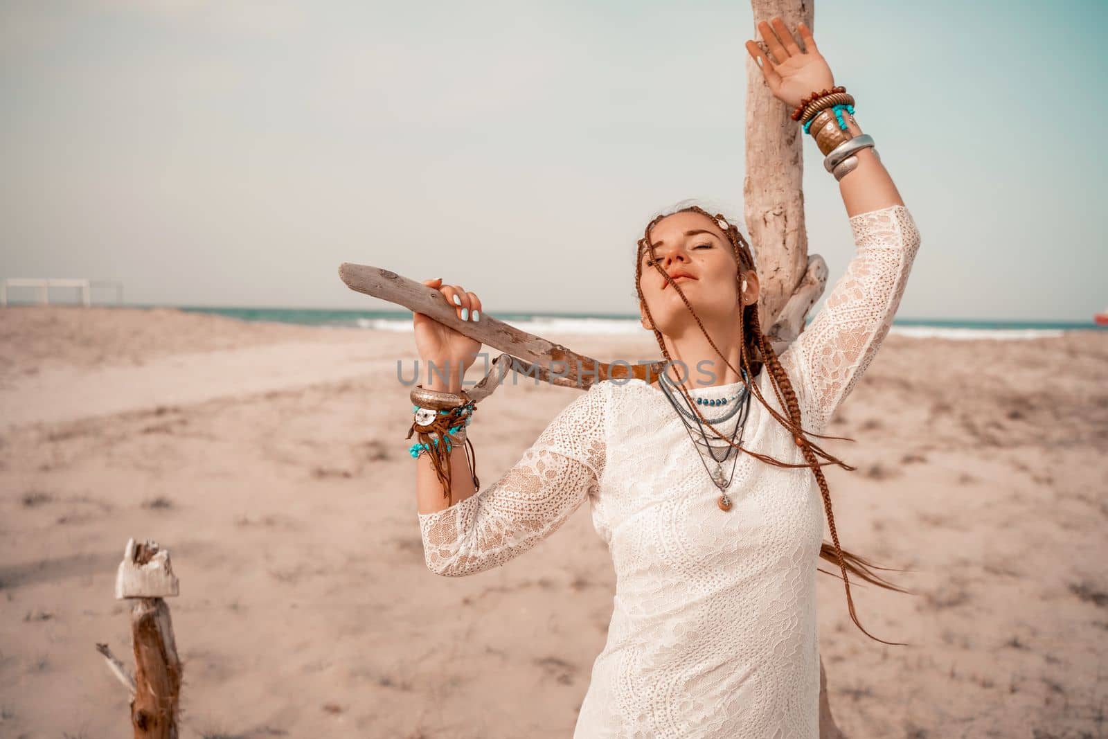 Model in boho style in a white long dress and silver jewelry on the beach. Her hair is braided, and there are many bracelets on her arms