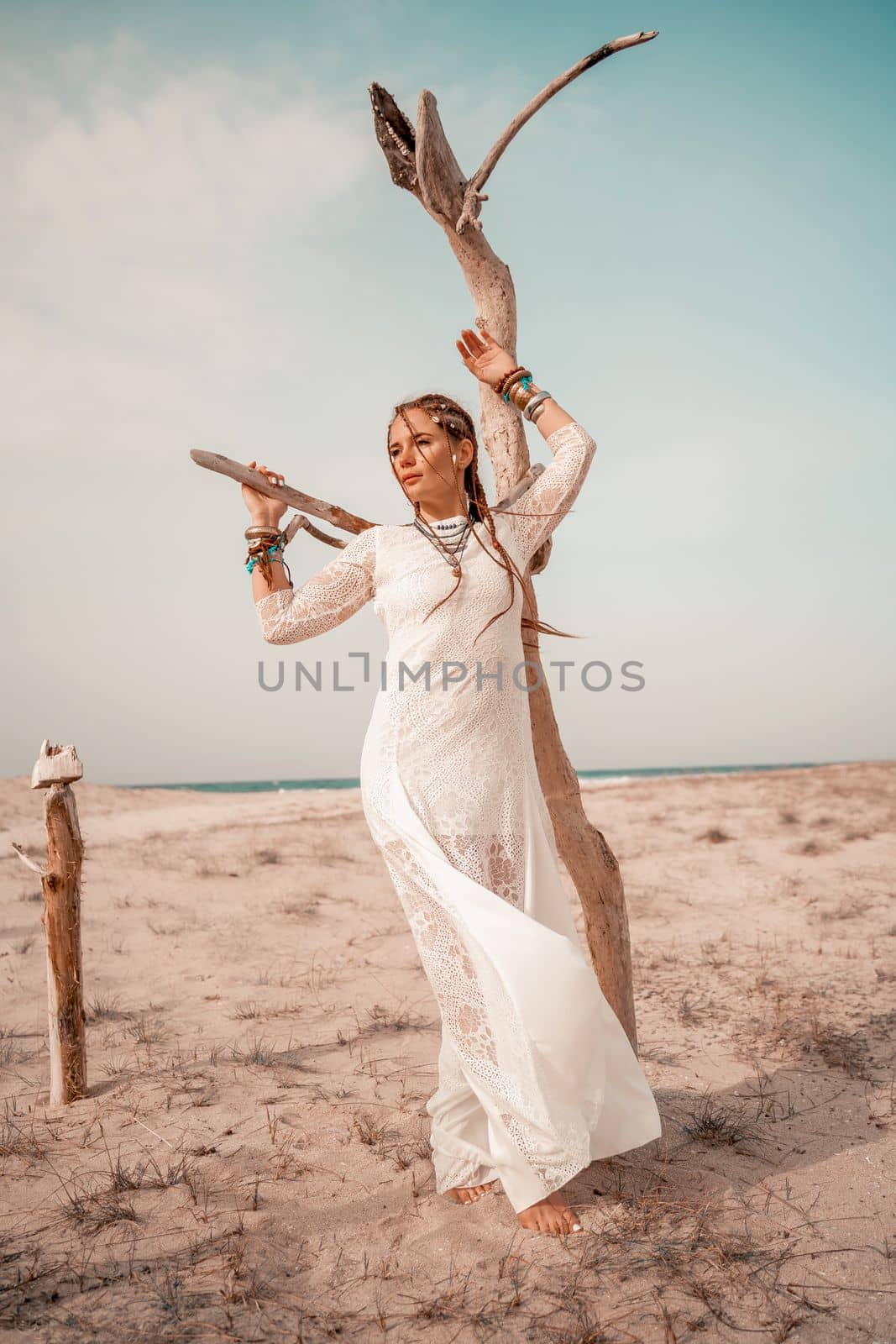 Model in boho style in a white long dress and silver jewelry on the beach. Her hair is braided, and there are many bracelets on her arms