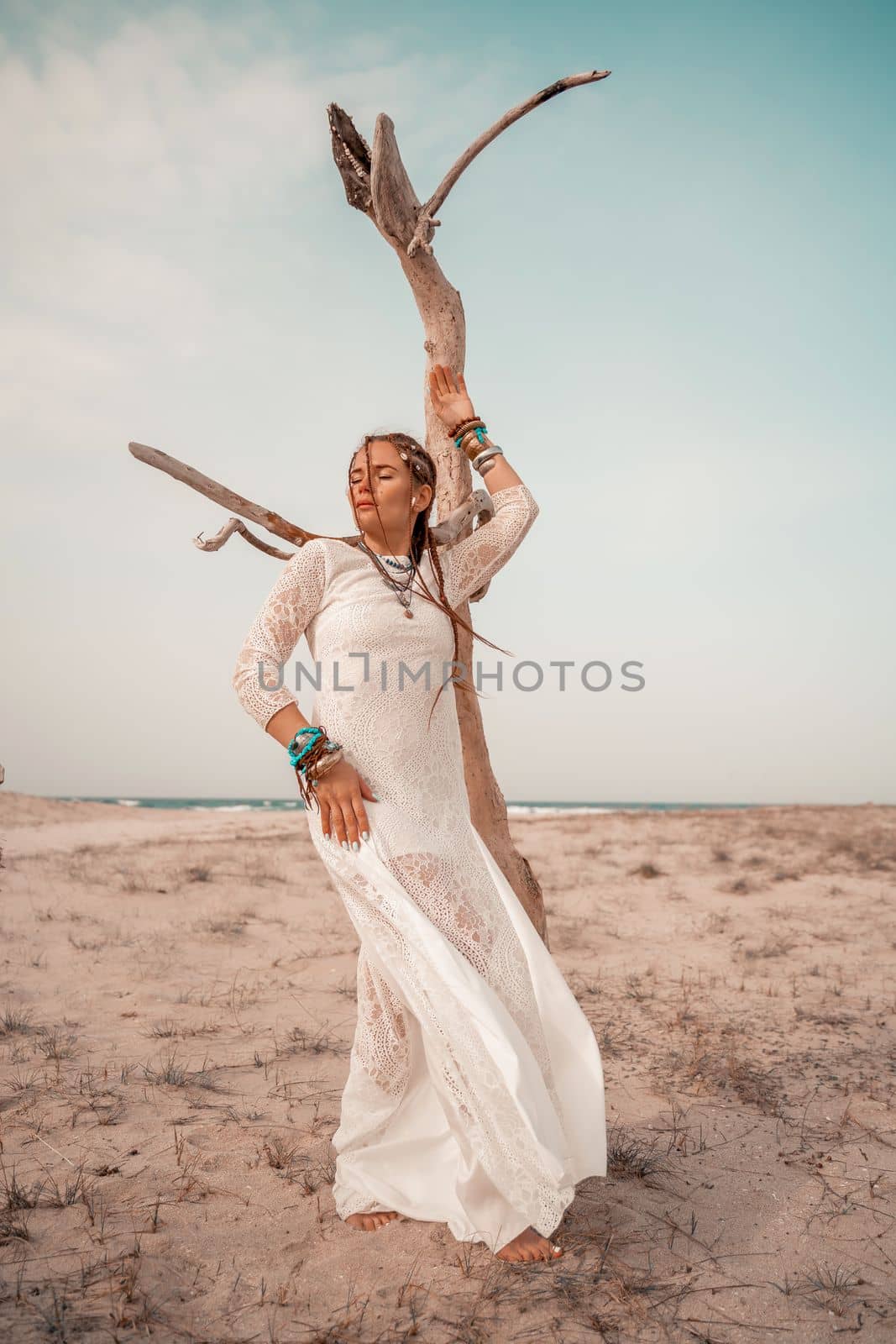 Model in boho style in a white long dress and silver jewelry on the beach. Her hair is braided, and there are many bracelets on her arms