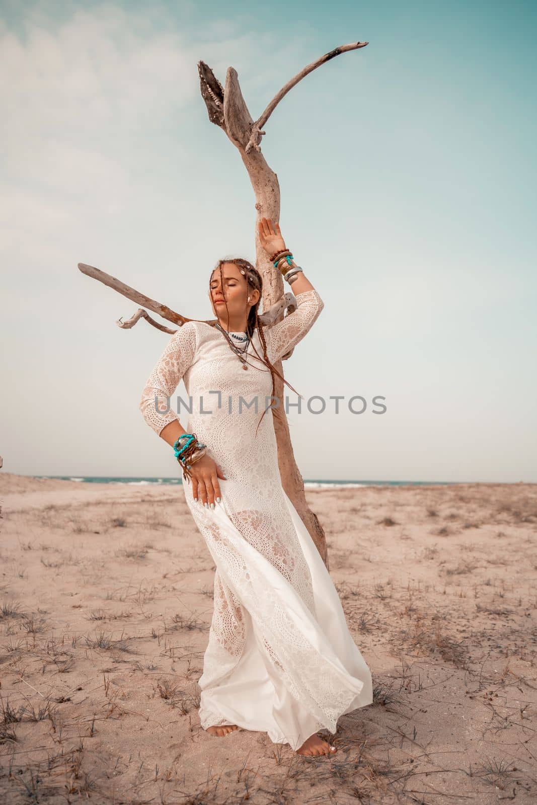 Model in boho style in a white long dress and silver jewelry on the beach. Her hair is braided, and there are many bracelets on her arms