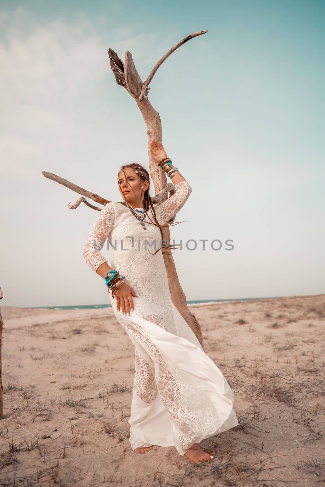 Model in boho style in a white long dress and silver jewelry on the beach. Her hair is braided, and there are many bracelets on her arms