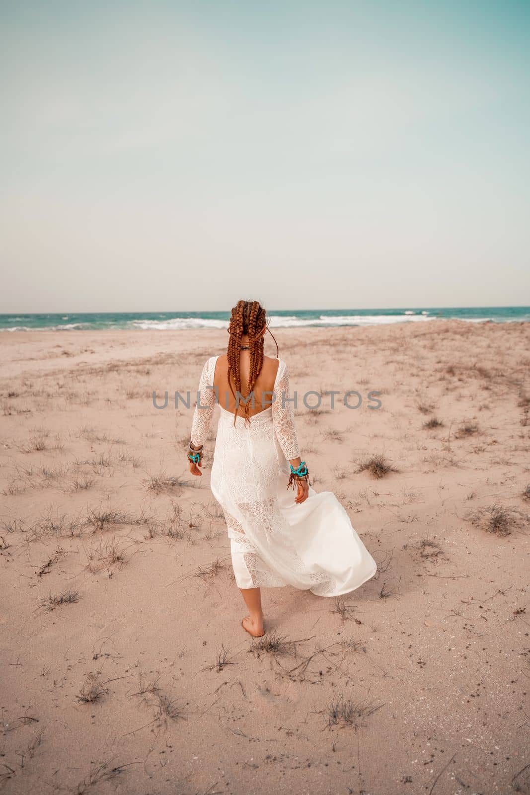 Model in boho style in a white long dress and silver jewelry on the beach. Her hair is braided, and there are many bracelets on her arms