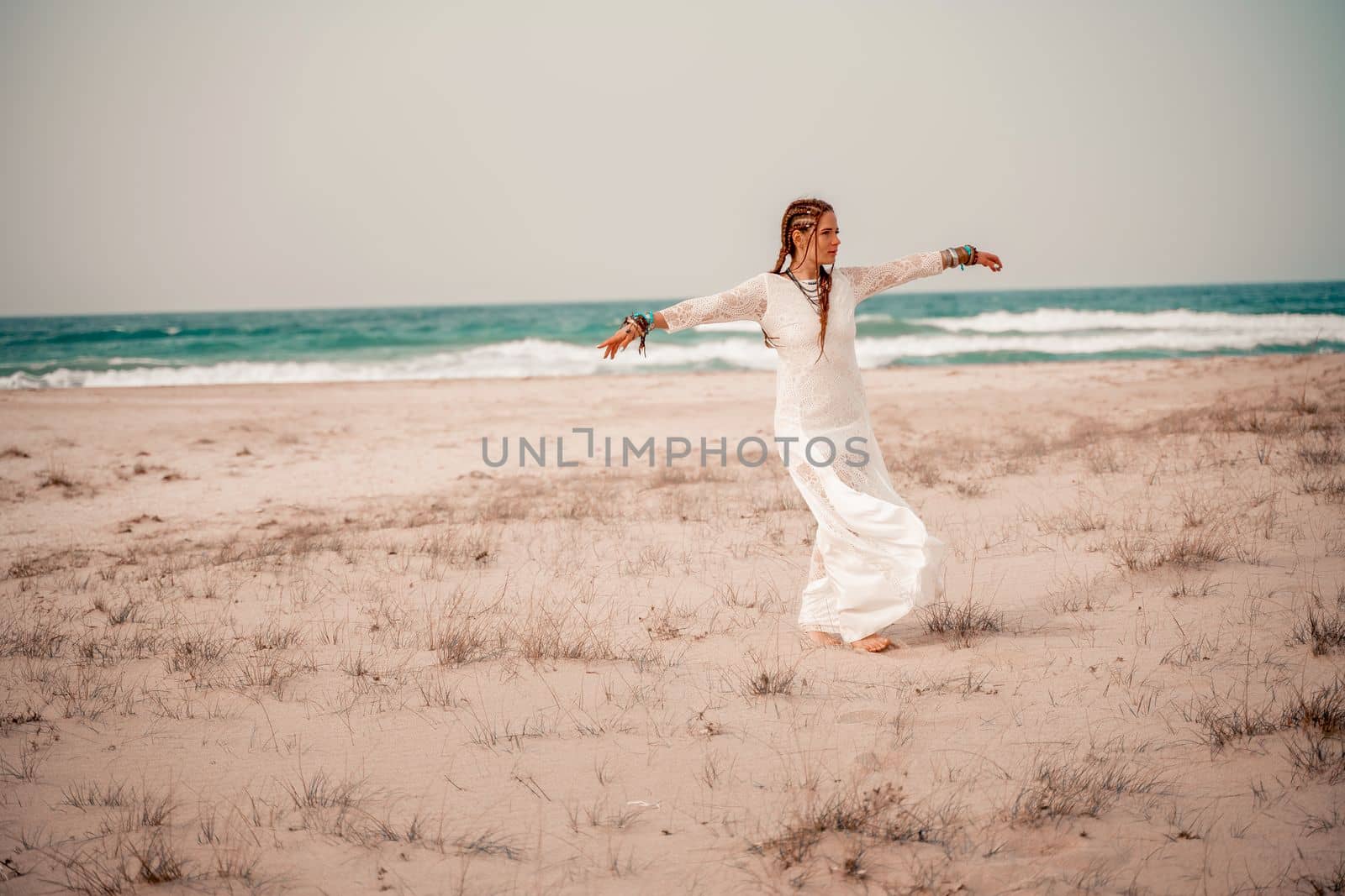 Model in boho style in a white long dress and silver jewelry on the beach. Her hair is braided, and there are many bracelets on her arms