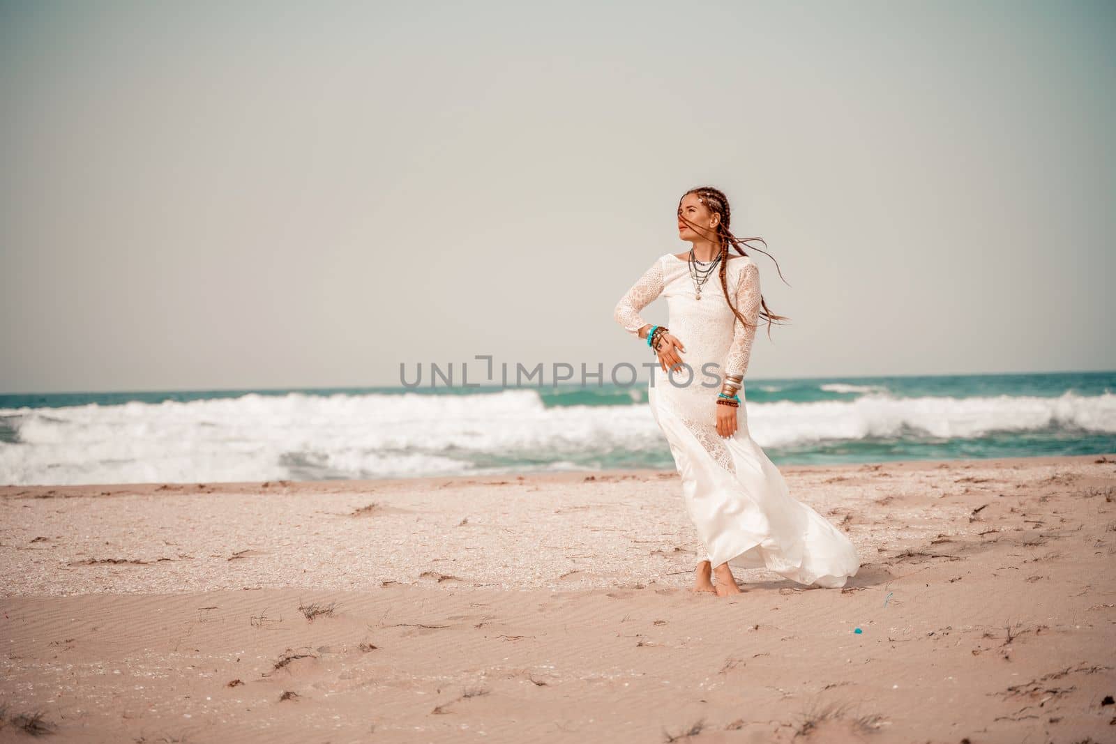 Model in boho style in a white long dress and silver jewelry on the beach. Her hair is braided, and there are many bracelets on her arms