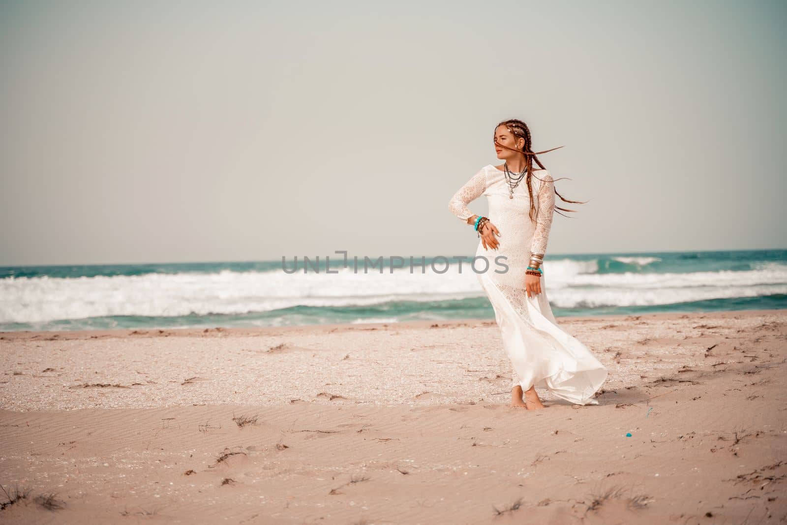 Model in boho style in a white long dress and silver jewelry on the beach. Her hair is braided, and there are many bracelets on her arms