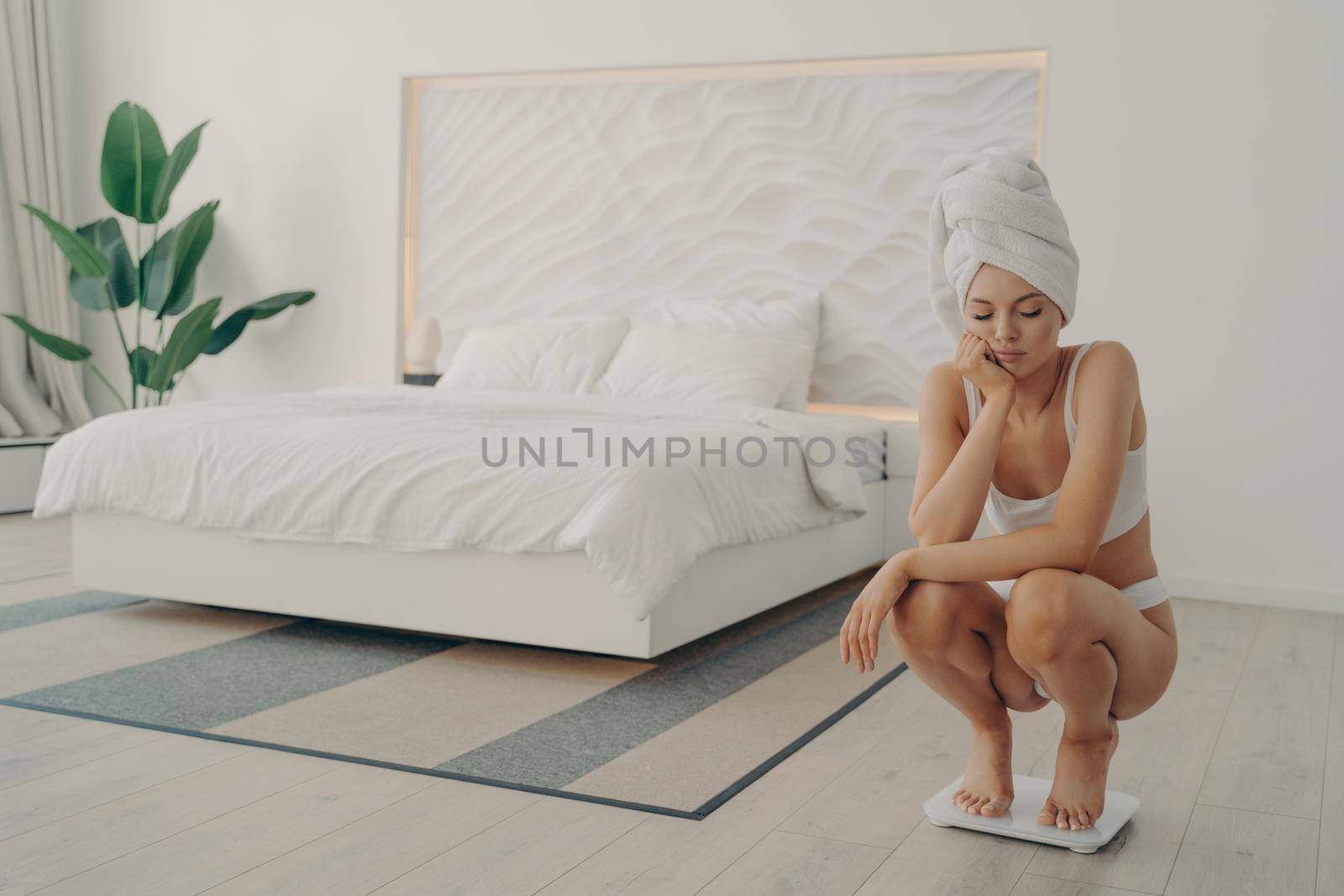 Dissatisfied young shapely woman crouched on scales in stylish light colored bedroom by vkstock