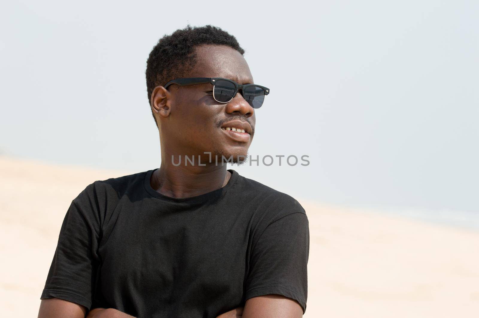 portrait of young man in black t-shirt standing arms crossed with black glasses and smiling at the beach.