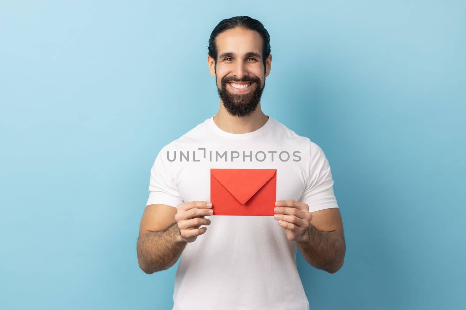 I got love letter on Valentine's day. Portrait of man with beard in white T-shirt holding letter in red envelope or greeting card and smiling joyfully. Indoor studio shot isolated on blue background.