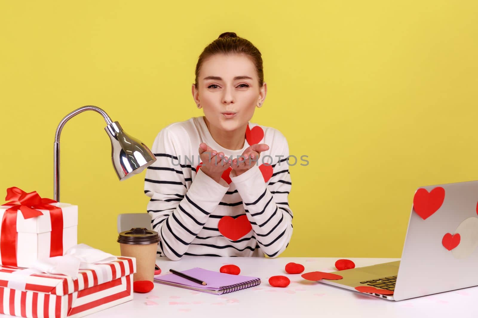 Woman sitting at workplace office, all covered with sticker love hearts, sending romantic sensual air kiss, full of valentine's day greetings. Indoor studio studio shot isolated on yellow background.