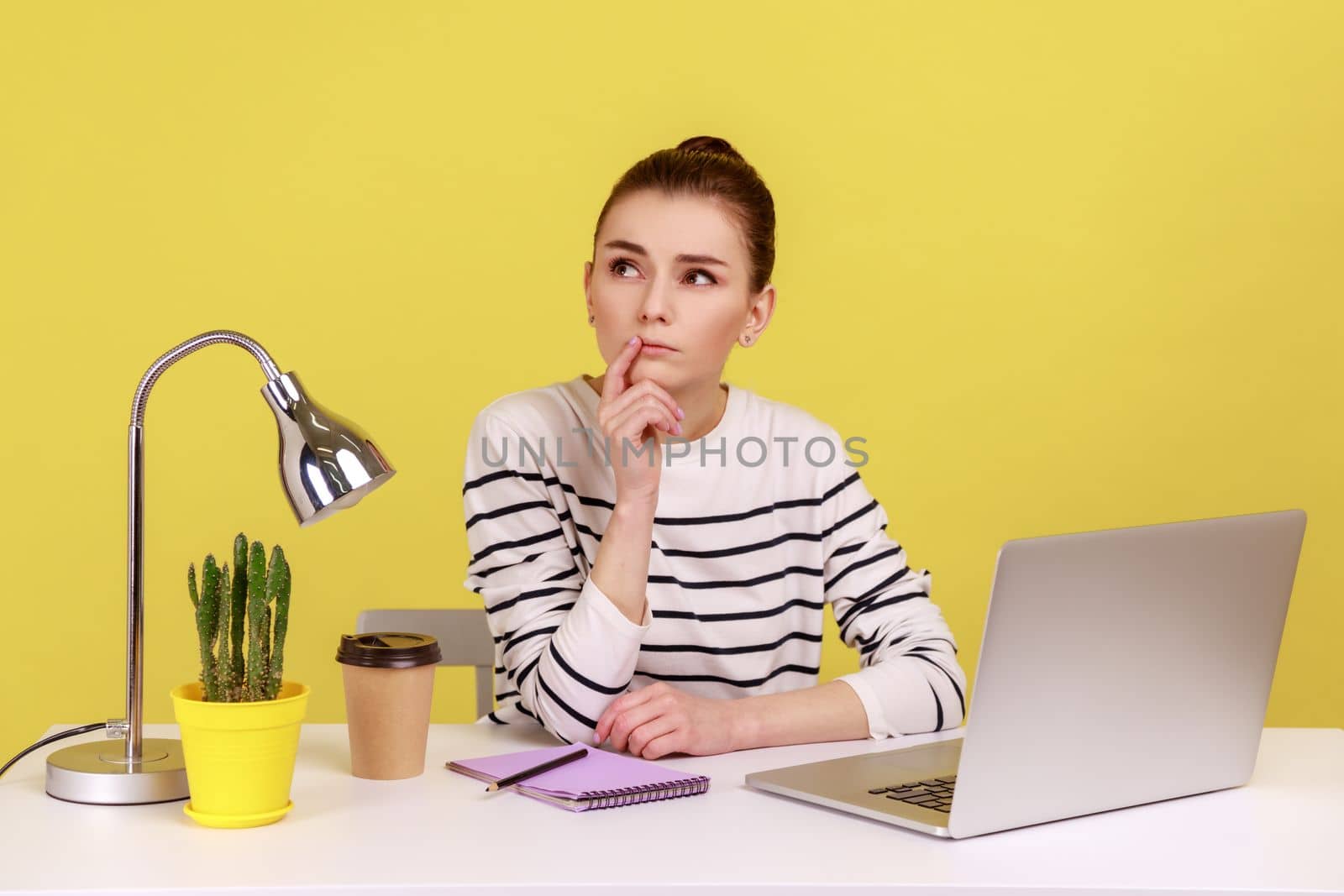 Pensive woman in striped shirt sitting with thoughtful confused expression while working on laptop in office, thinking over new project. Indoor studio studio shot isolated on yellow background.