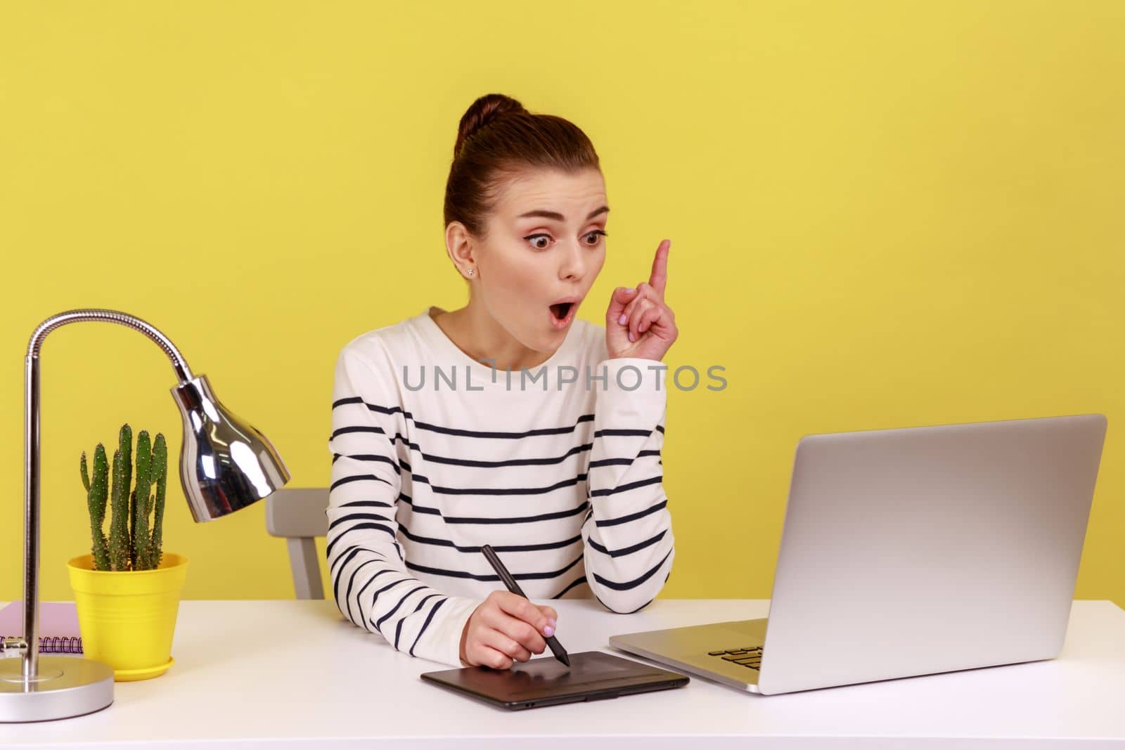 Excited woman designer pointing finger up, having idea, drawing with stylus on graphic tablet, sitting in office working on new collection. Indoor studio studio shot isolated on yellow background.