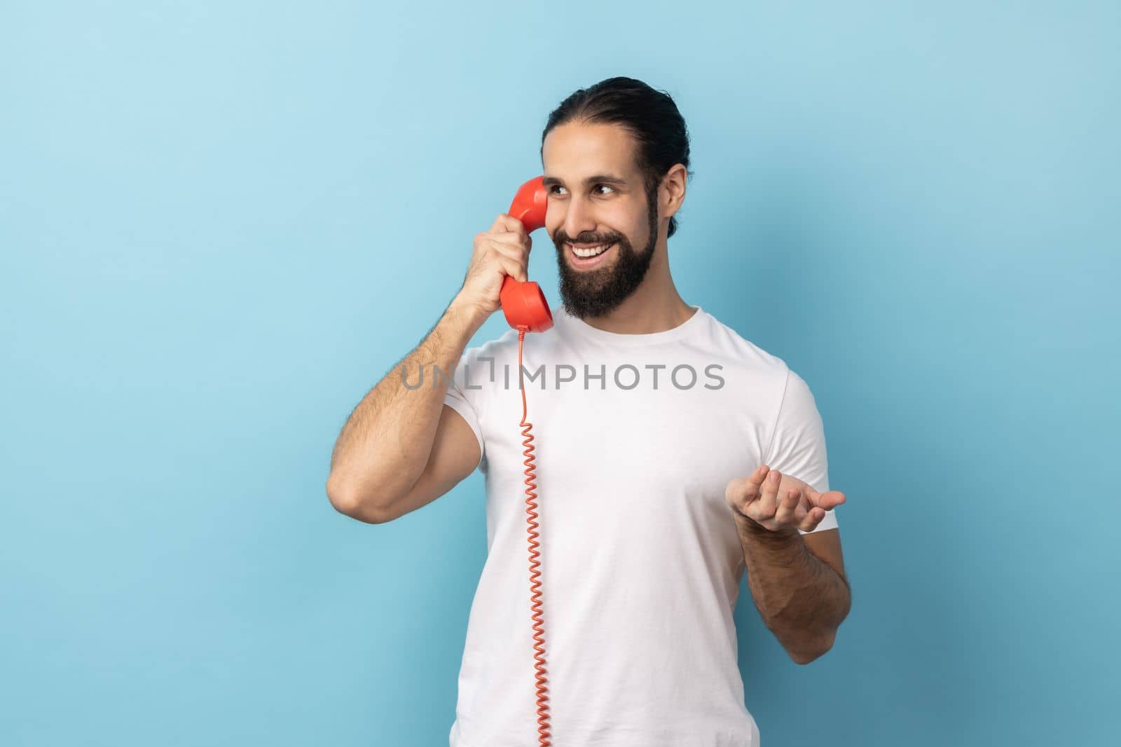Portrait of positive man with beard wearing white T-shirt talking on red retro telephone, having pleasant conversation, looking away with smile Indoor studio shot isolated on blue background.