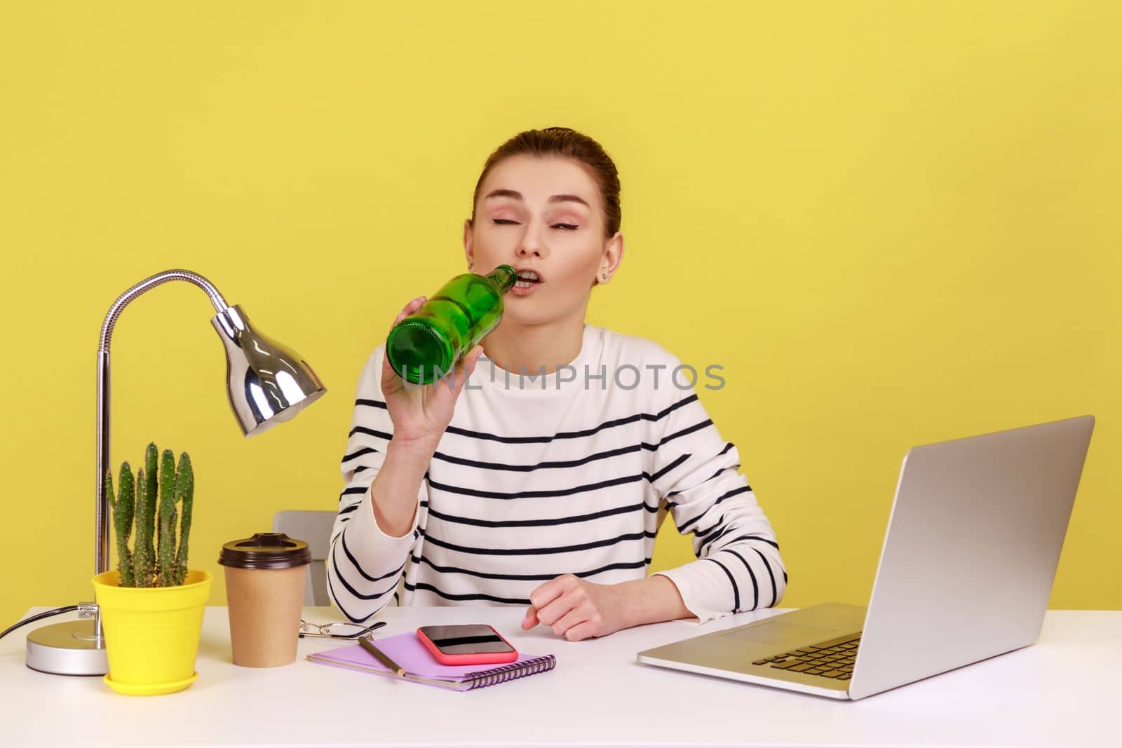 Portrait of young adult woman office manger drinking beer, sitting on workplace with closed eyes, being drunk during working day. Indoor studio studio shot isolated on yellow background.
