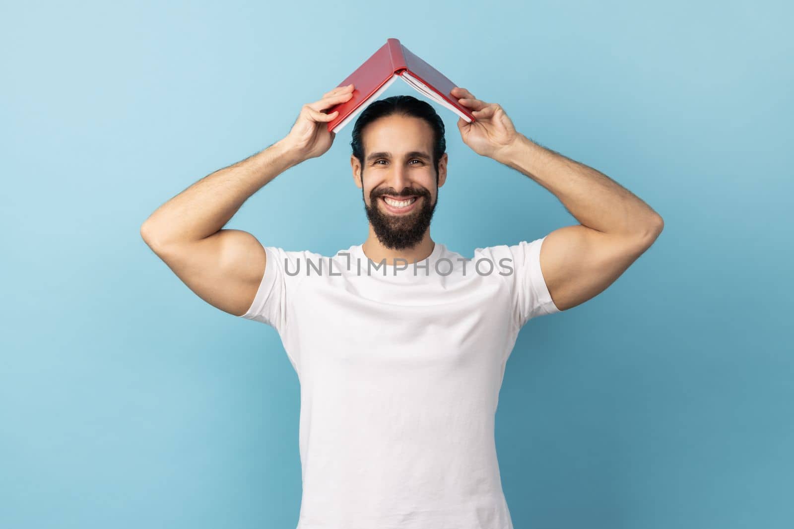 Portrait of delighted man with beard wearing white T-shirt making roof, holding book over head, looking at camera with happiness. Indoor studio shot isolated on blue background.