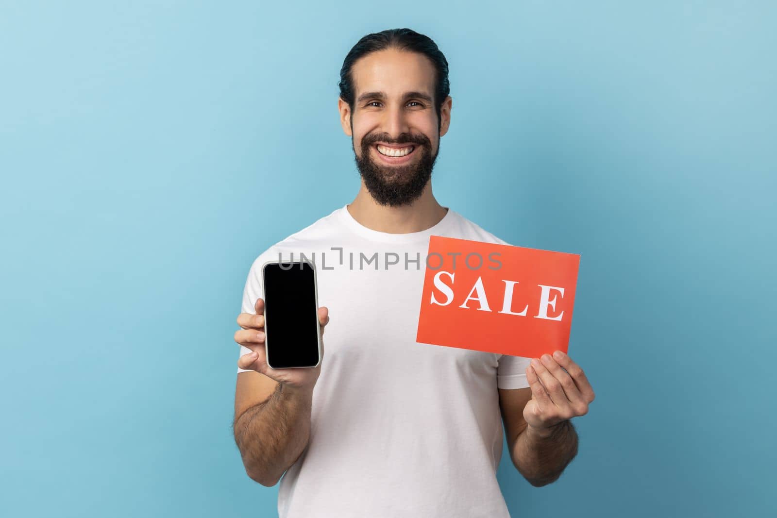 Portrait of handsome smiling man with beard wearing white T-shirt holding sale card and smartphone, being satisfied with online sell-out. Indoor studio shot isolated on blue background.